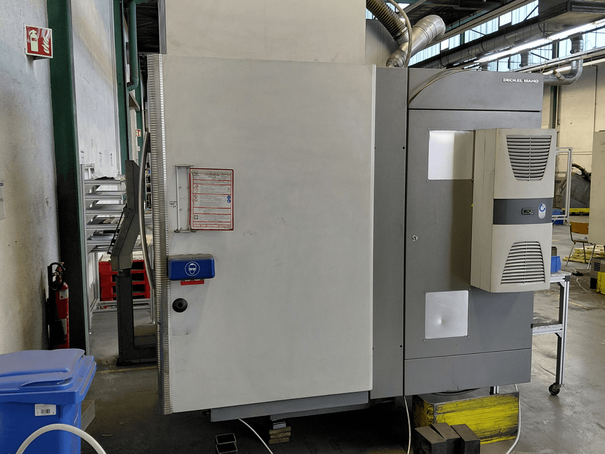 Vista laterale del centro di lavoro verticale DECKEL MAHO DMC 635 V con pannello di controllo ed etichette di sicurezza visibili.