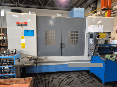 Macchina Mazak VTC-300C in uno stabilimento, vista frontale che mostra il pannello di controllo, le porte grigie con griglia di finestre e le attrezzature vicine.