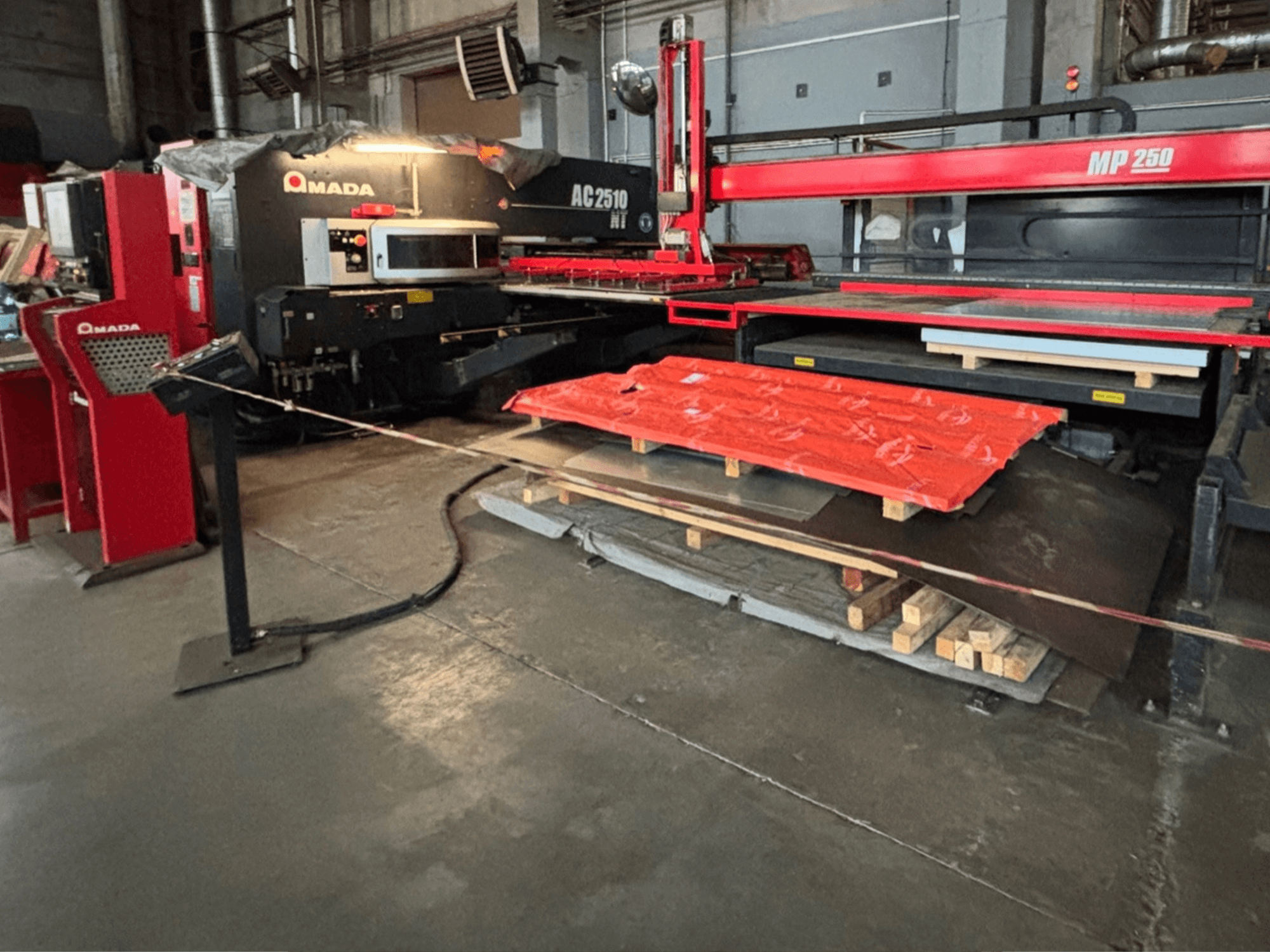 Taglierina laser Amada AC2510 NT in fabbrica, vista frontale, macchinari pesanti con lamiere e pallet sul pavimento.