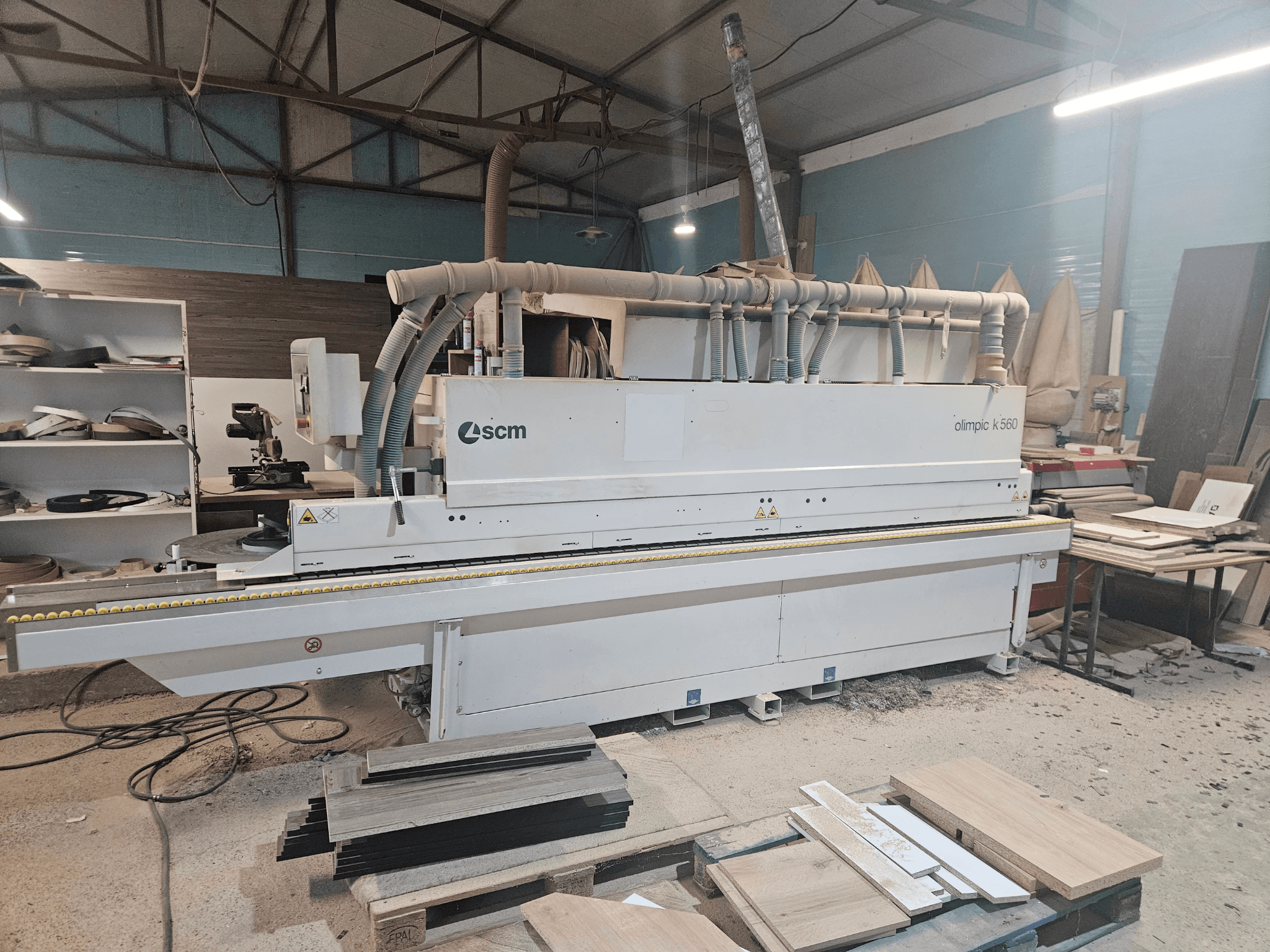 Macchina bordatrice SCM Olimpic K560 in un'officina, vista frontale, con pannelli di legno e utensili visibili sul pavimento.