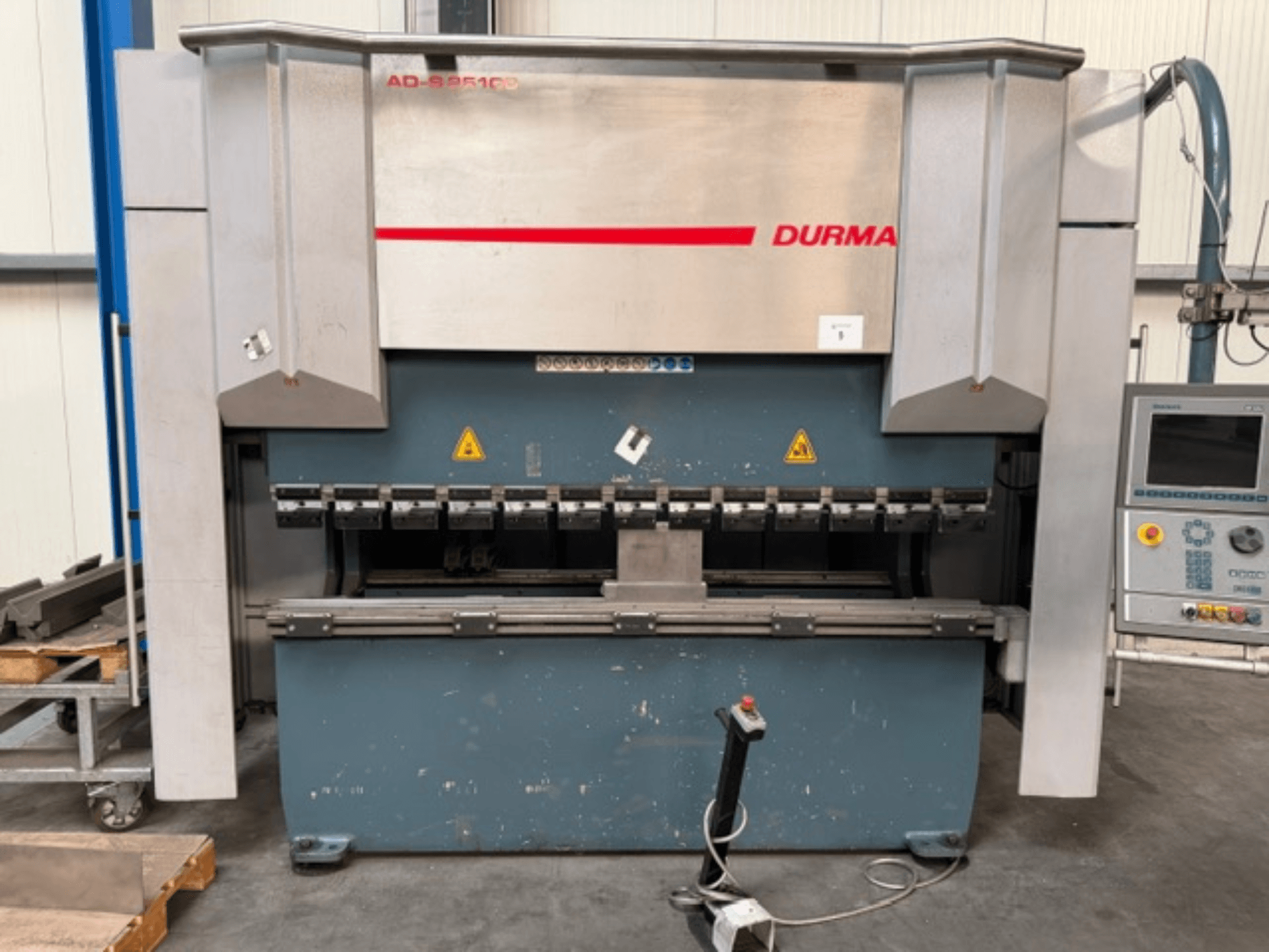 Vista frontale della pressa piegatrice Durma ADS 25100 con pannello di controllo, che mostra gli utensili di piegatura e le etichette di sicurezza.