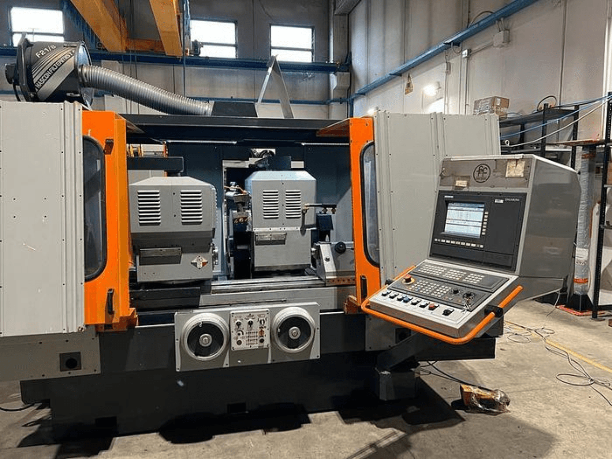 Vista frontale di una macchina CNC con doppio mandrino, pannello di controllo e accenti arancioni. Presenta un condotto di ventilazione e controlli operativi.