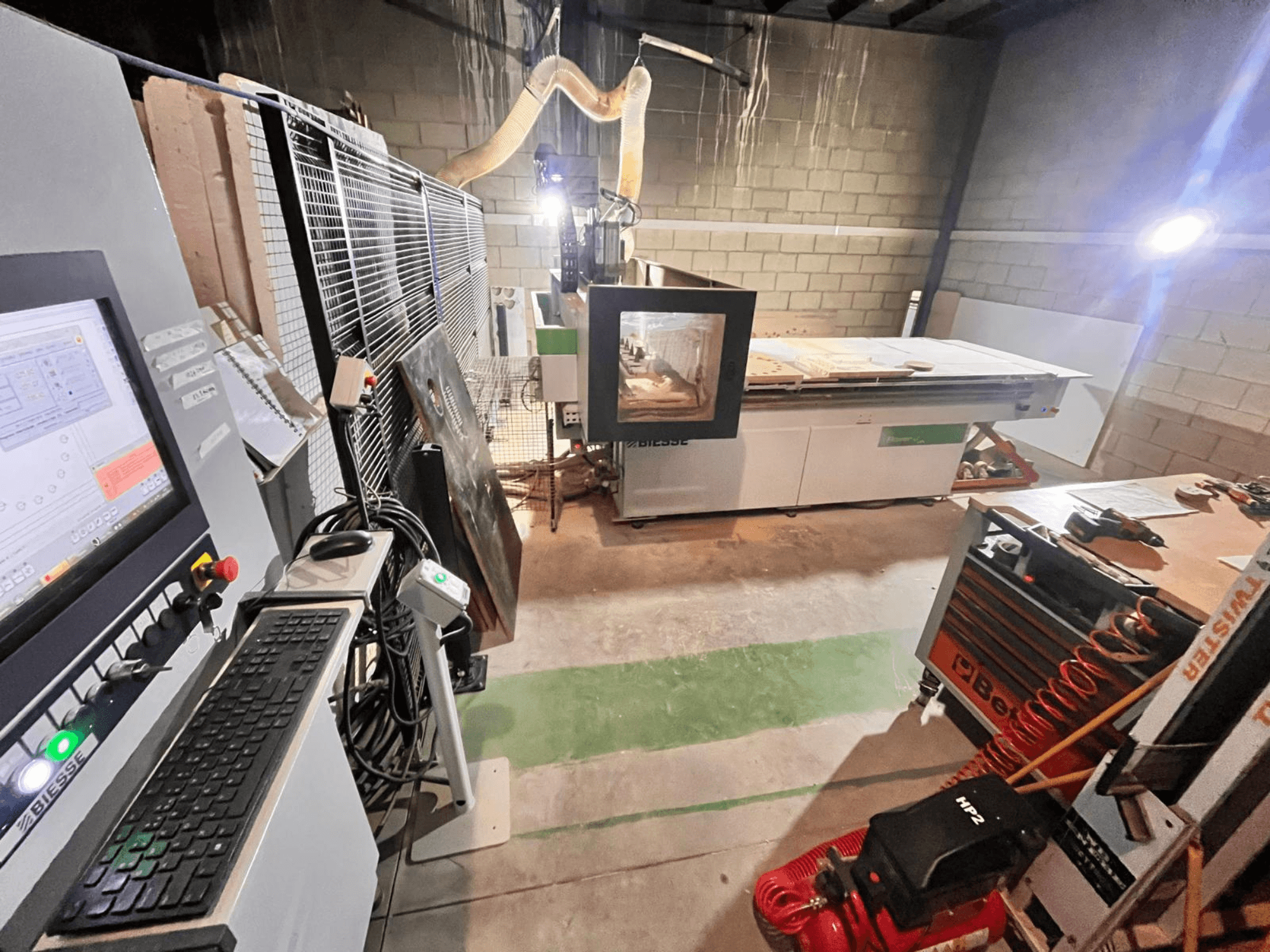 Macchina CNC Biesse in un'officina, vista laterale che mostra il pannello di controllo, l'aspirazione della polvere, gli utensili e i vari materiali sul tavolo.