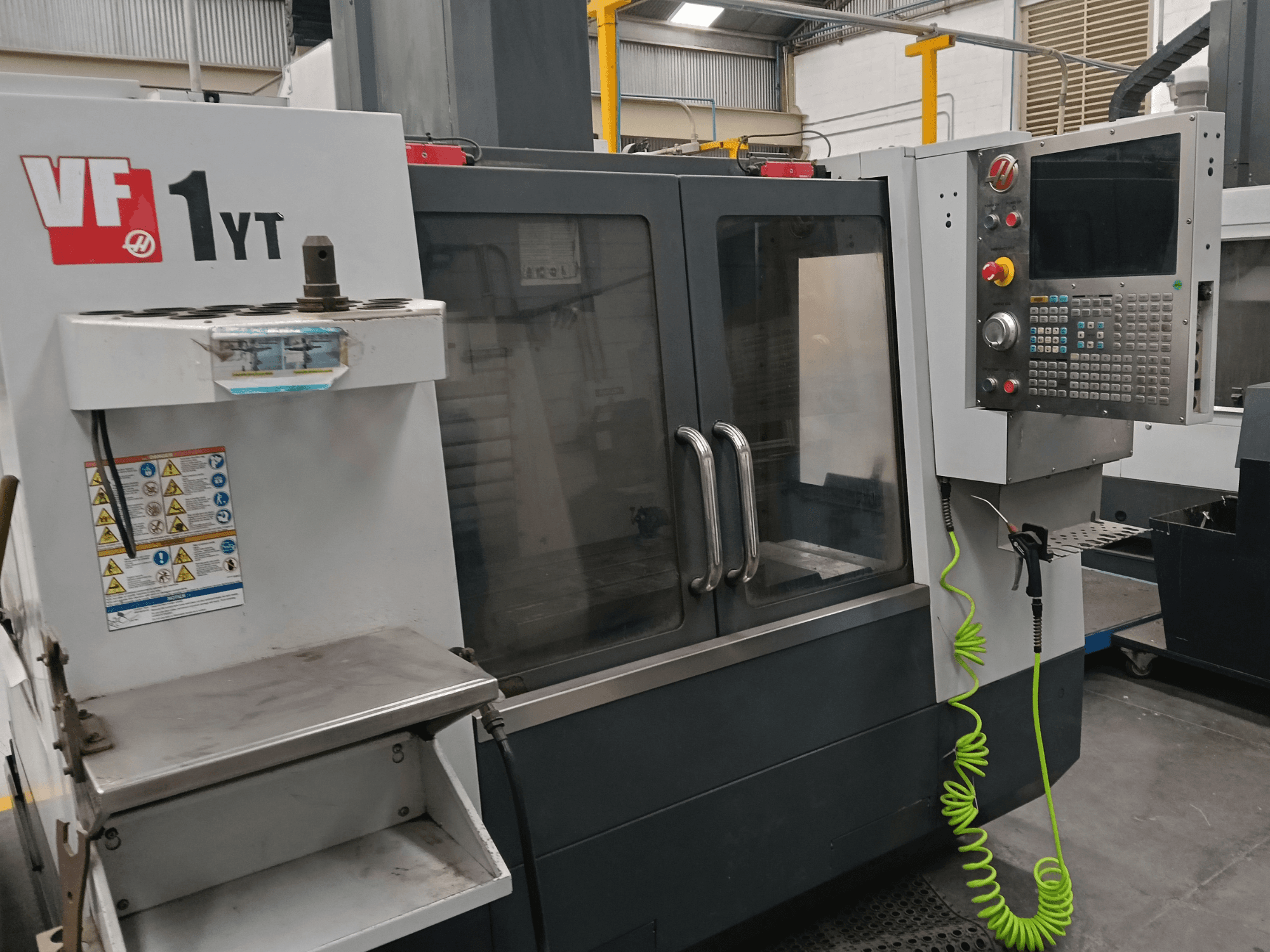 Centro di lavorazione verticale HAAS VF-1YT-2, vista frontale, con pannello di controllo, portautensili, etichette di sicurezza e area di lavorazione.