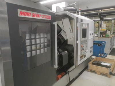 Macchina CNC MORI SEIKI NZ1500 con una leggera angolazione, che mostra il pannello di controllo e l'area di lavoro con pezzi metallici su un pallet di legno.
