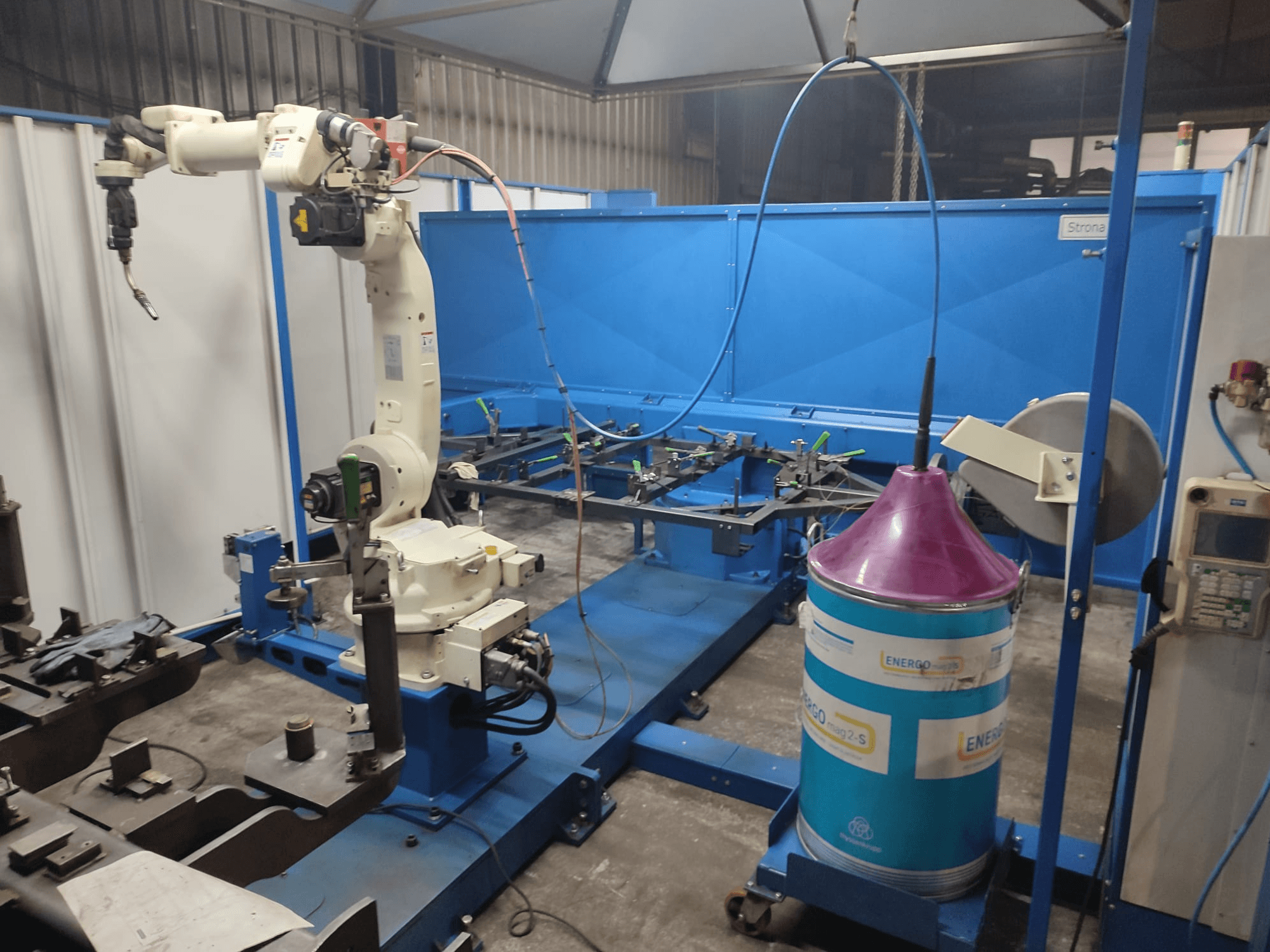 Braccio robotico industriale con base bianca e blu, allestito in un'officina con una superficie di lavoro blu e componenti metallici.