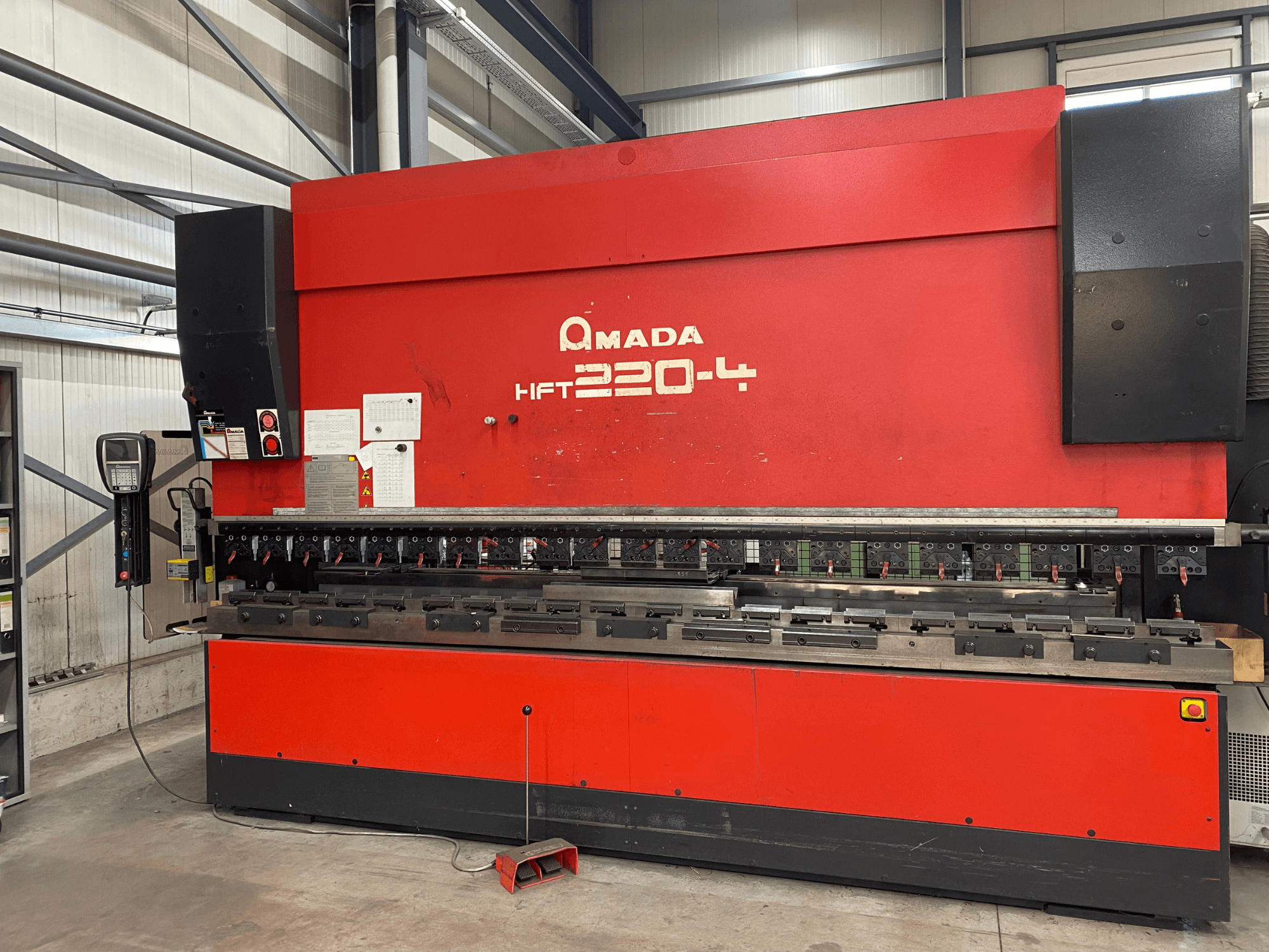 Pressa piegatrice AMADA HFT 220-4 rossa in un ambiente di fabbrica, vista frontale che mostra il pannello di controllo e gli utensili di piegatura sul piano di lavoro.