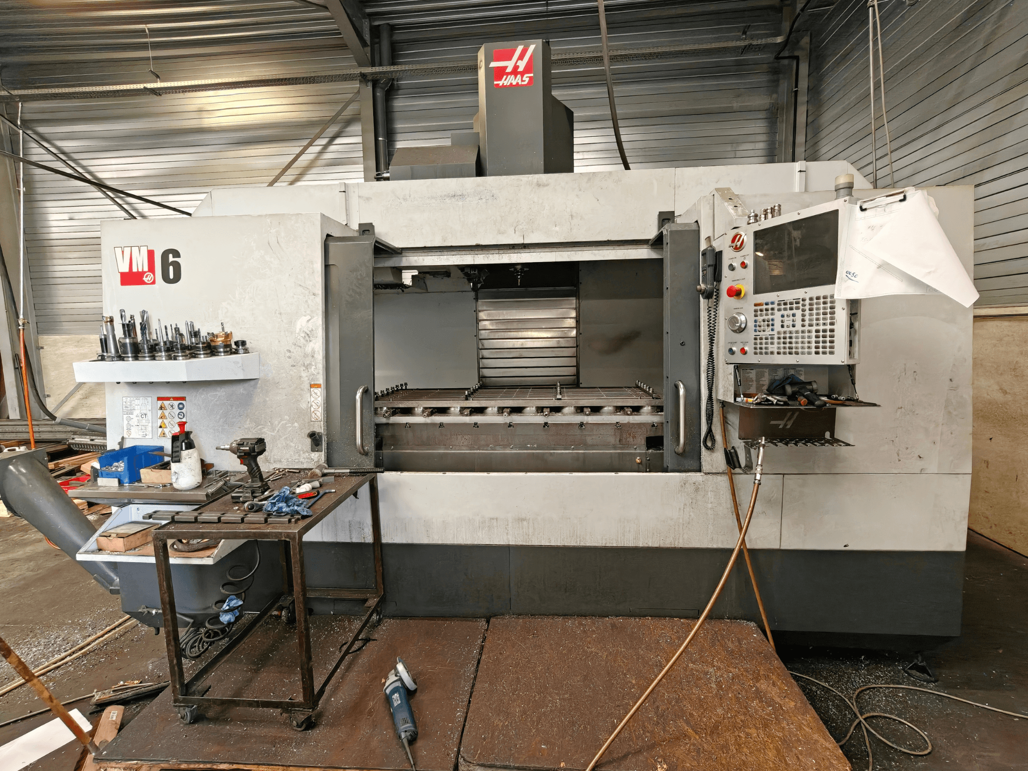 Centro di lavorazione verticale Haas VM-6, vista frontale, con magazzino utensili, pannello di controllo e detriti nell'area di lavoro visibili.