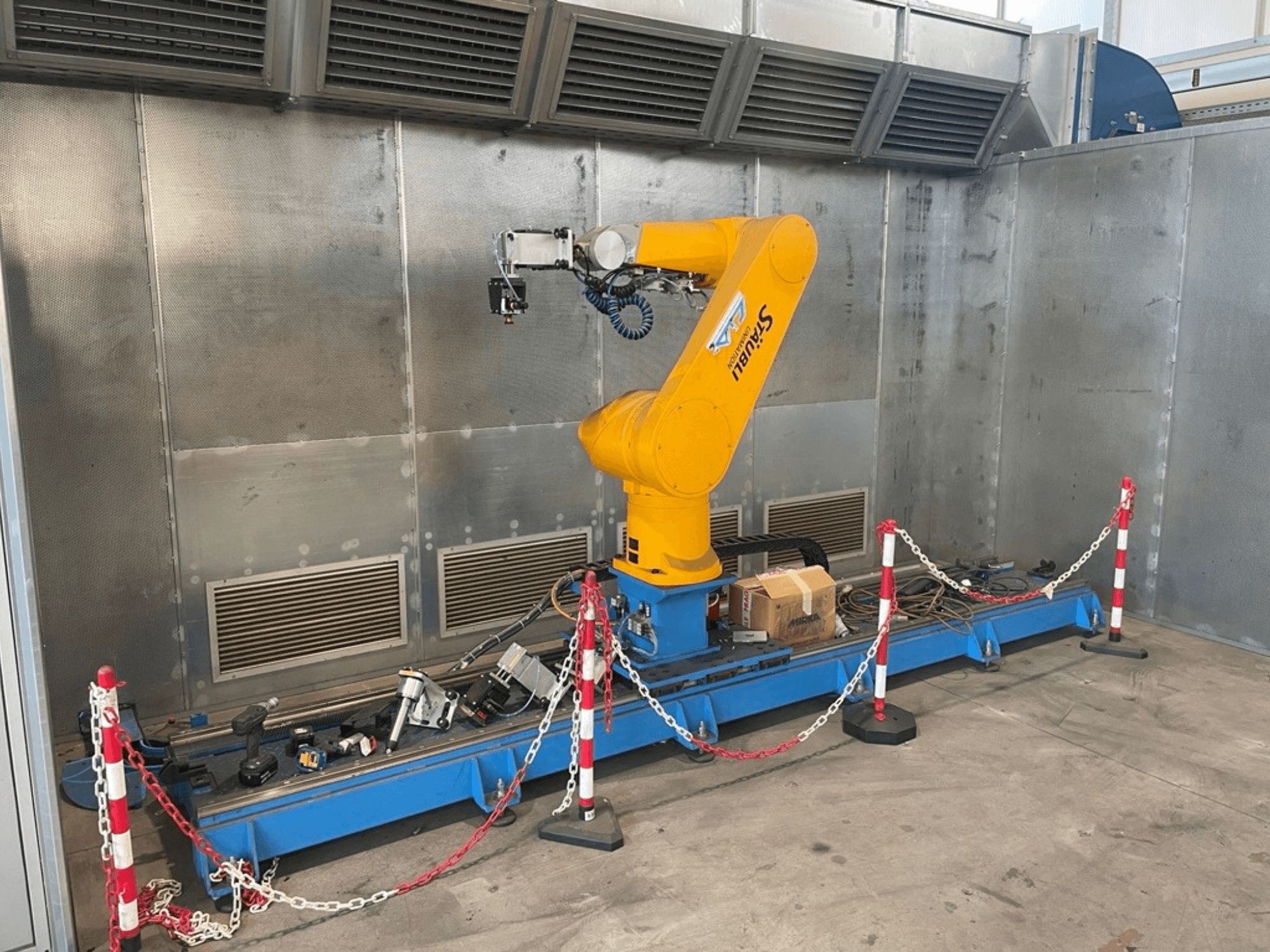 Braccio robotico STAUBLI CS8C-RX160 in un'officina, vista laterale, con cavi e vari strumenti su una base blu circondata da barriere protettive.