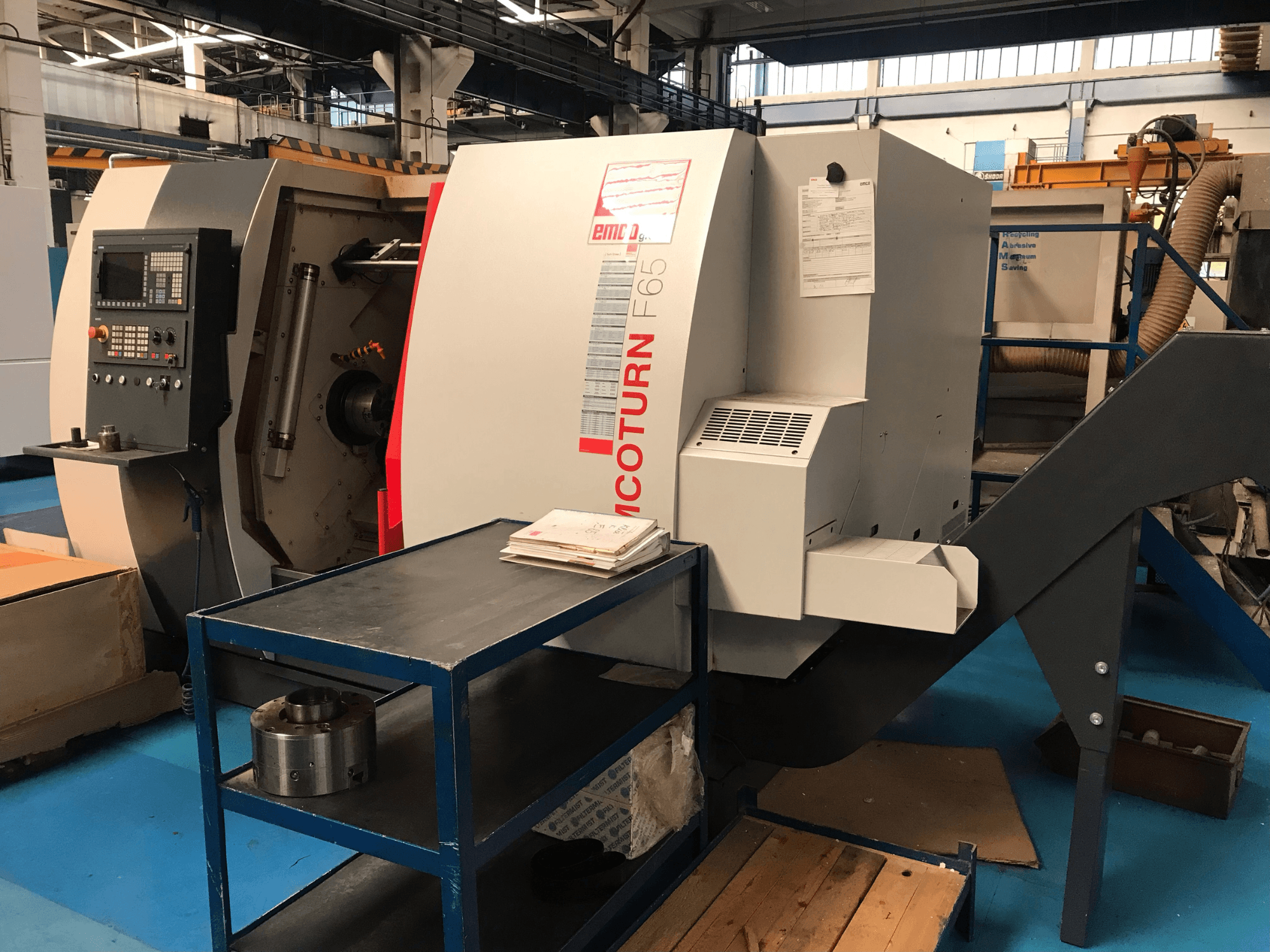 Tornio orizzontale EMCO Emcoturn 65 visto di profilo, con i comandi e l'area di lavoro in primo piano e alcuni componenti metallici nelle vicinanze.
