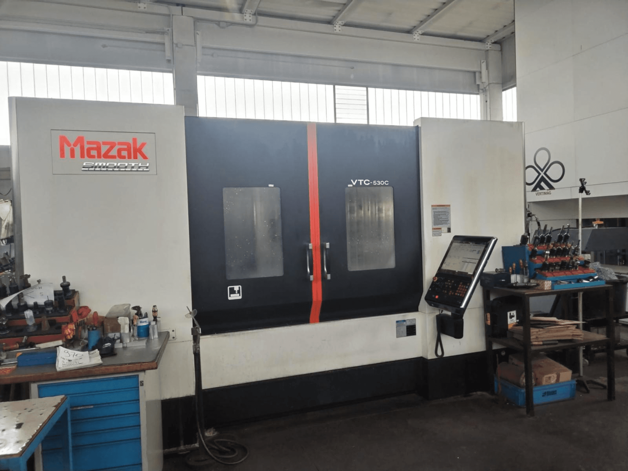 Centro di lavorazione verticale Mazak VTC-530C in un'officina, con pannello di controllo e utensili su un tavolo laterale.