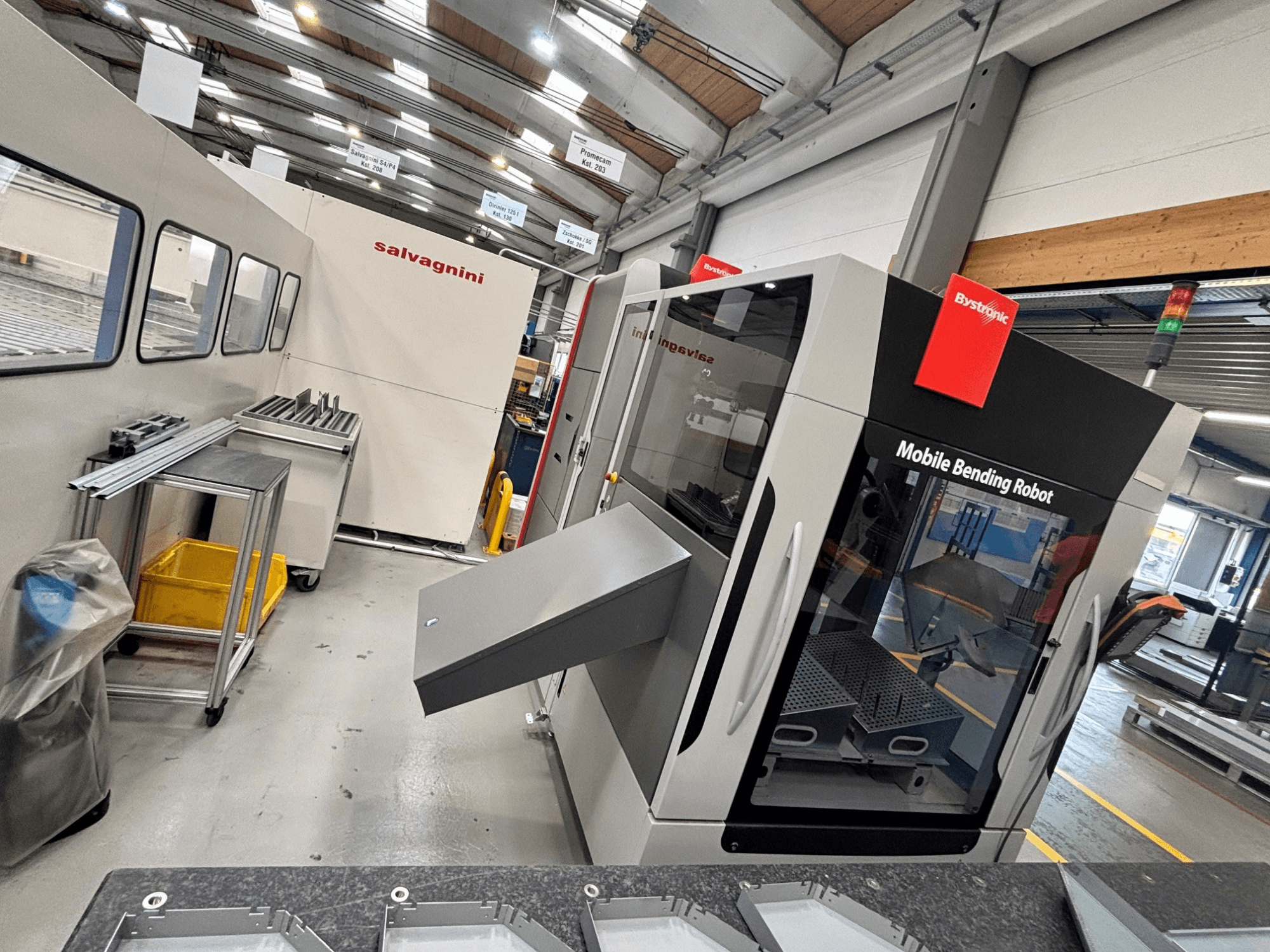 Pressa piegatrice Bystronic Xpert 40/1030, vista frontale che mostra il robot di piegatura mobile e l'area di lavoro con pezzi metallici su un tavolo.