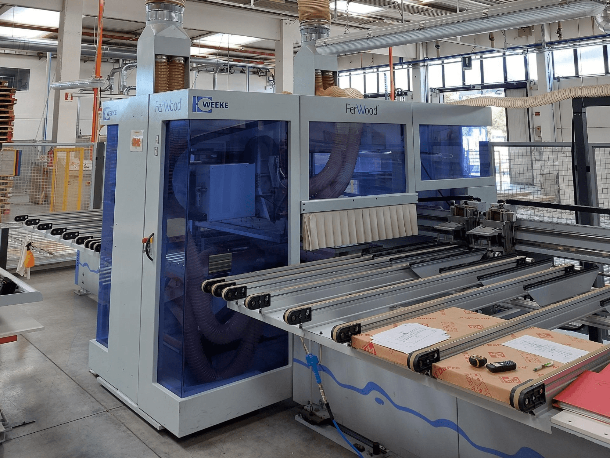 Macchina CNC Weeke FerWood in un'officina, vista frontale che mostra i pannelli colorati di blu, i pulsanti di controllo e il piano di lavoro con i materiali.