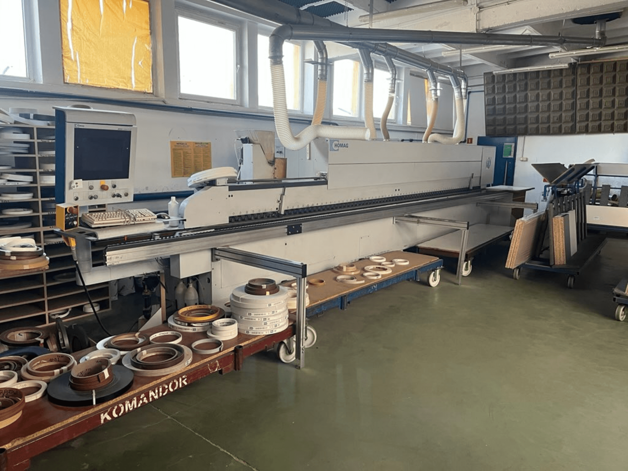 Macchina bordatrice HOMAG in un'officina, con comandi e vari materiali per la bordatura su un carrello blu. Vista laterale.
