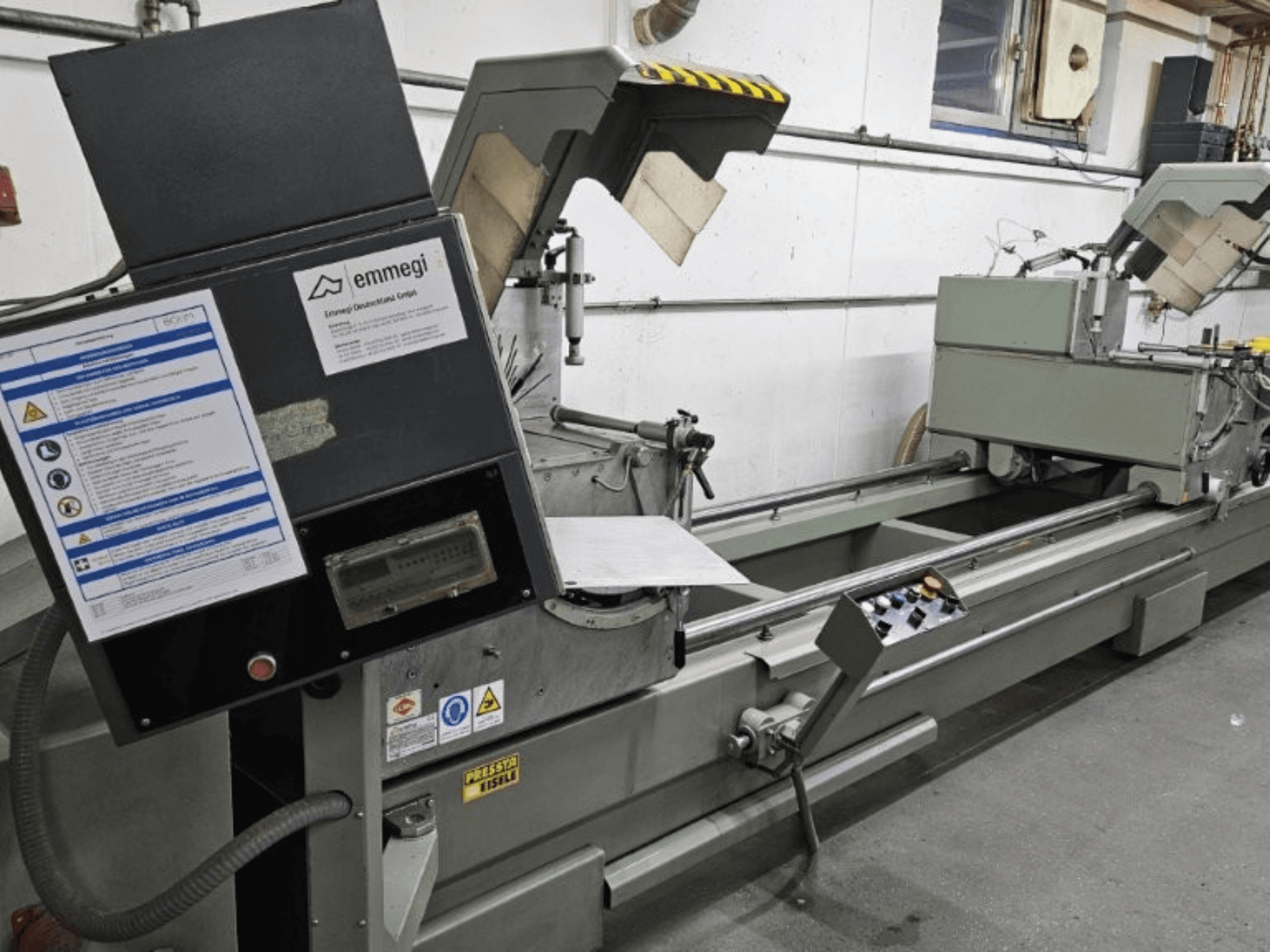 Macchina da taglio industriale Emmegi, vista frontale, con pannello di controllo, etichette di sicurezza e componenti operativi.