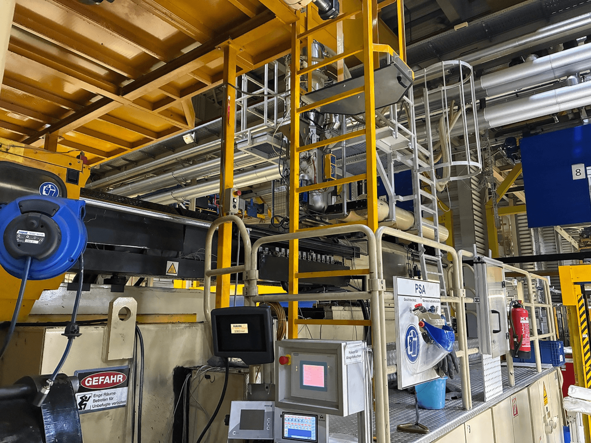 Pannello di controllo di un macchinario industriale con attrezzature di sicurezza blu e impalcature gialle, vista laterale con tubi e segnali di pericolo.