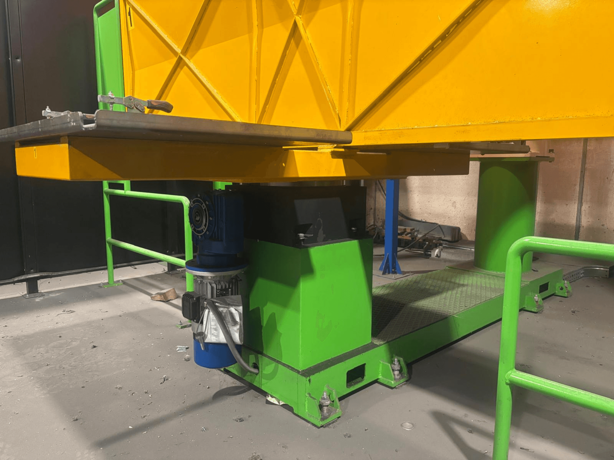 Macchinario industriale verde e giallo visto frontalmente, con motore e piattaforma con superficie strutturata.