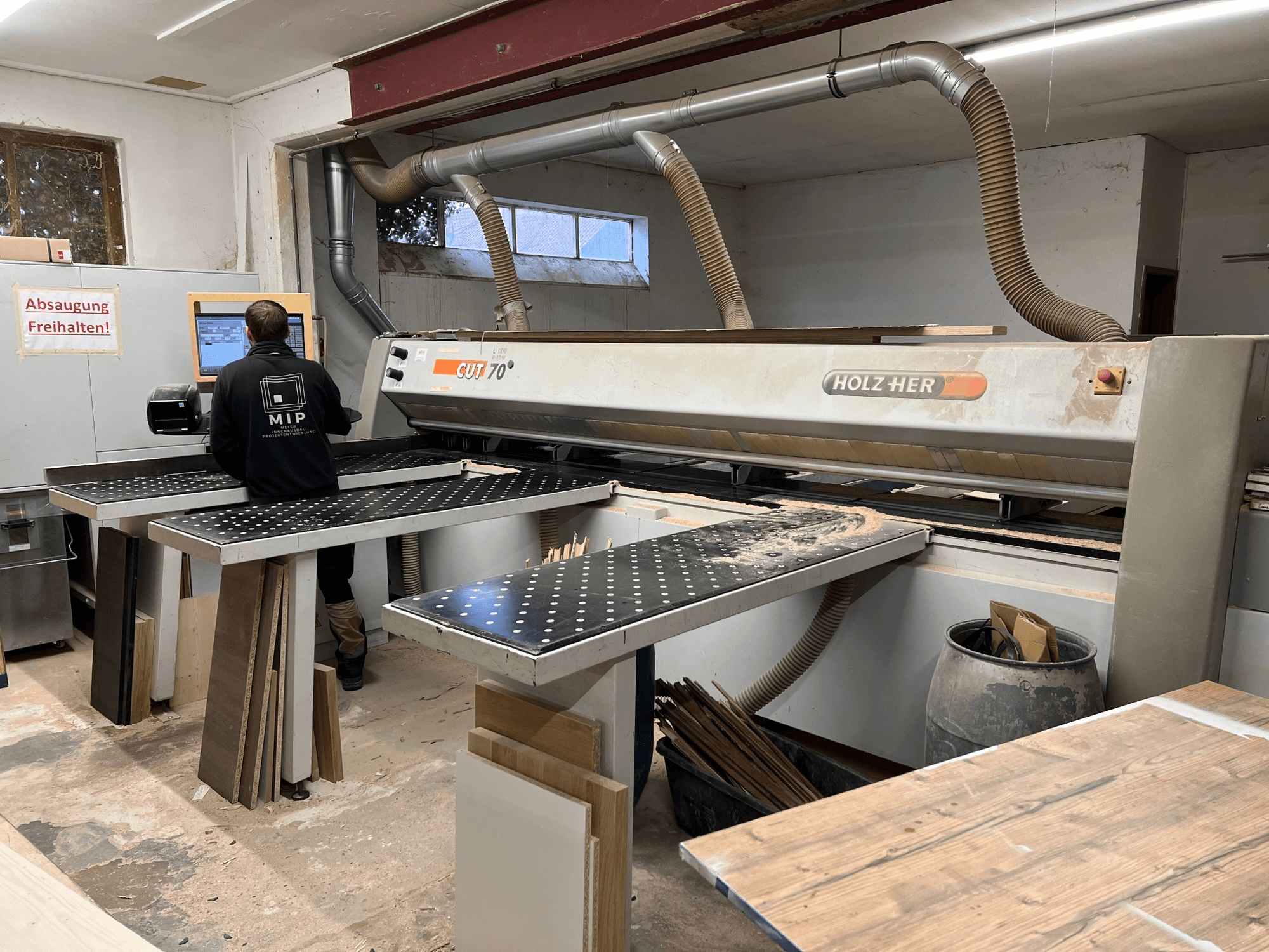 Macchina da taglio HOLZHER CUT 70 in officina; vista frontale che mostra l'operatore, i piani di lavoro e i materiali in legno impilati sulle superfici.