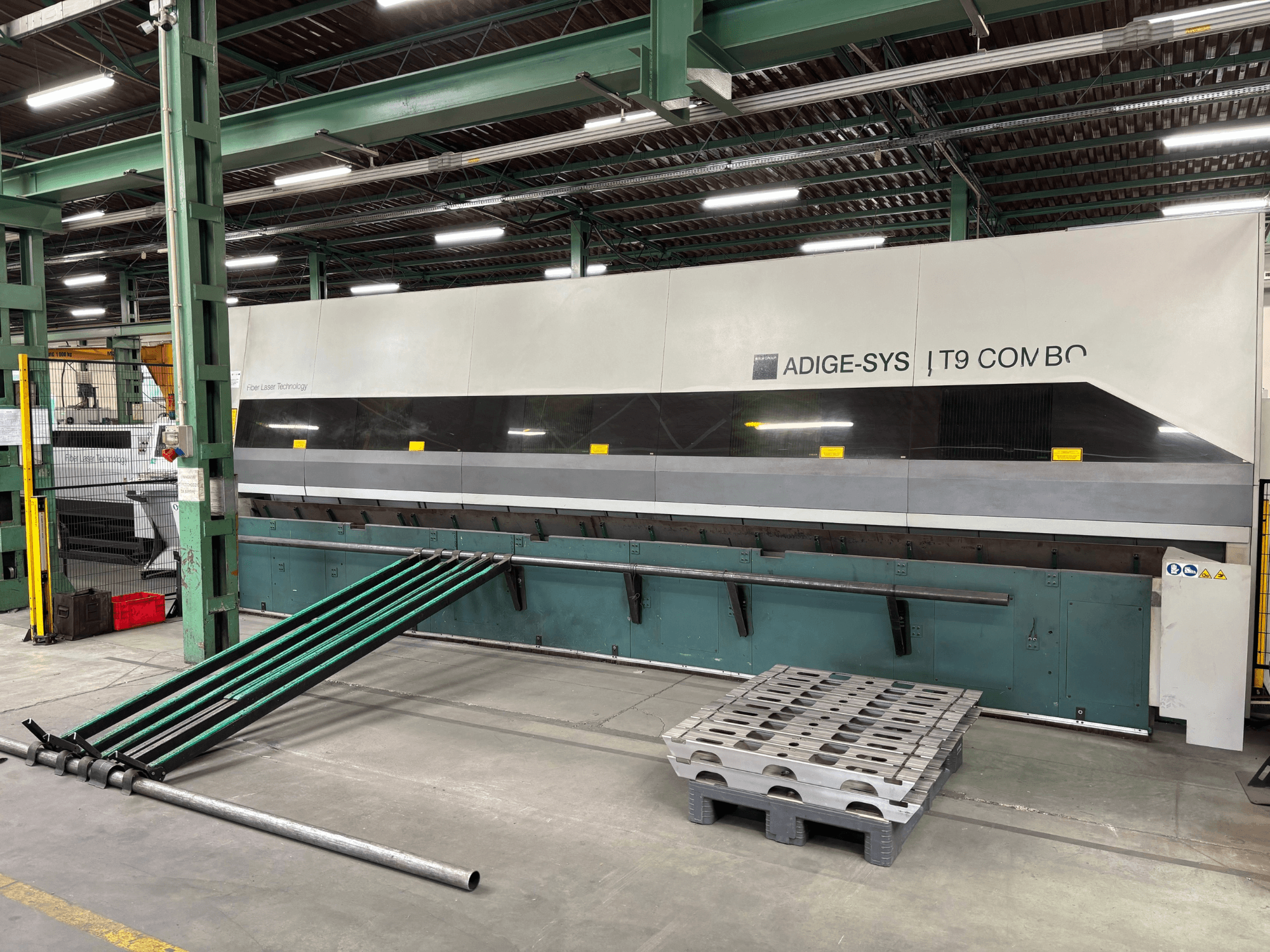 Vista frontale della macchina combinata per il taglio di tubi BLM Adige LT9 con bracci di carico verdi e pallet sul pavimento dello stabilimento.