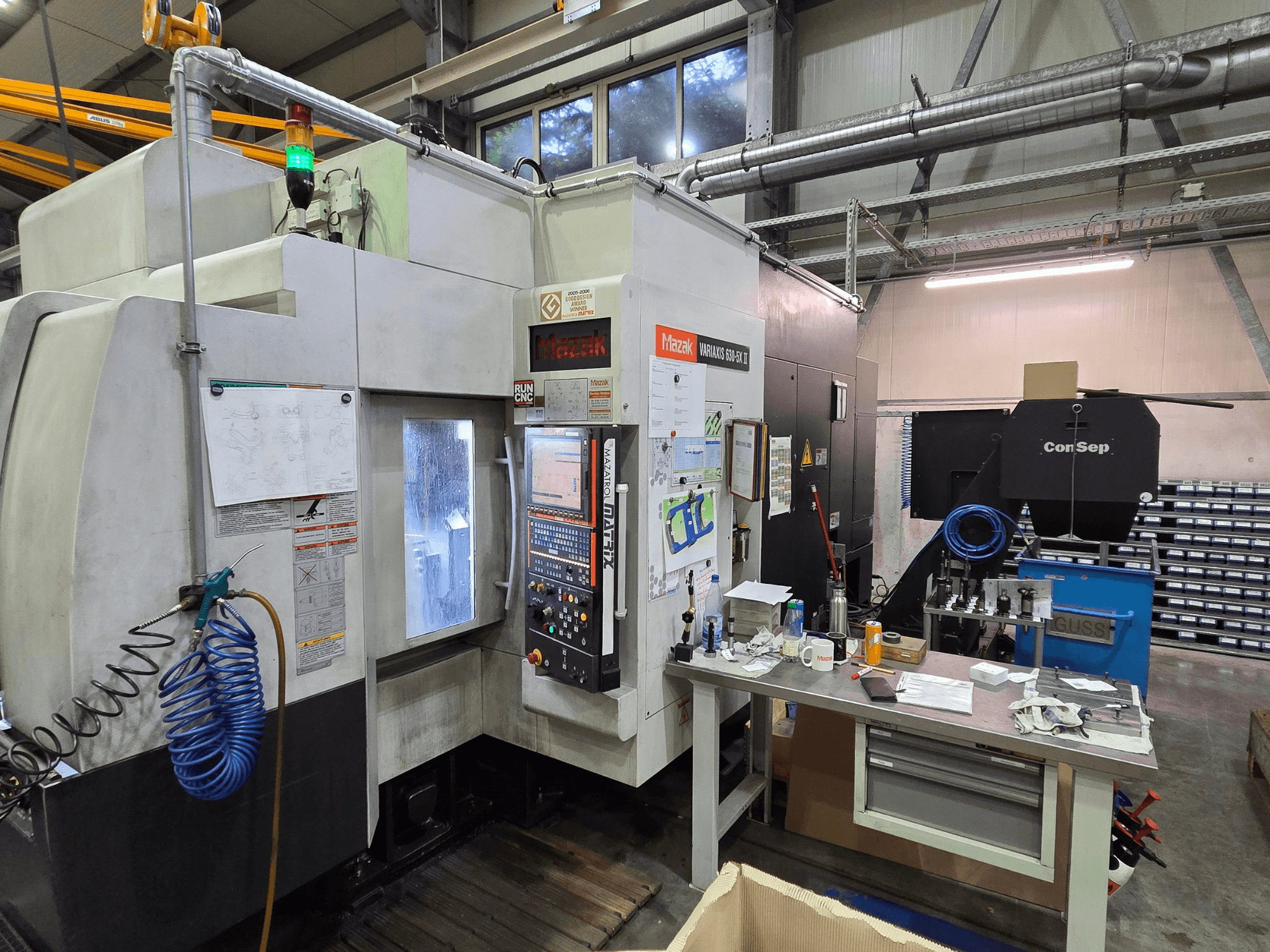 Macchina CNC Mazak Variaxis 630-5X in un'officina, vista frontale con pannello di controllo, tubo flessibile e accessori sul tavolo laterale.