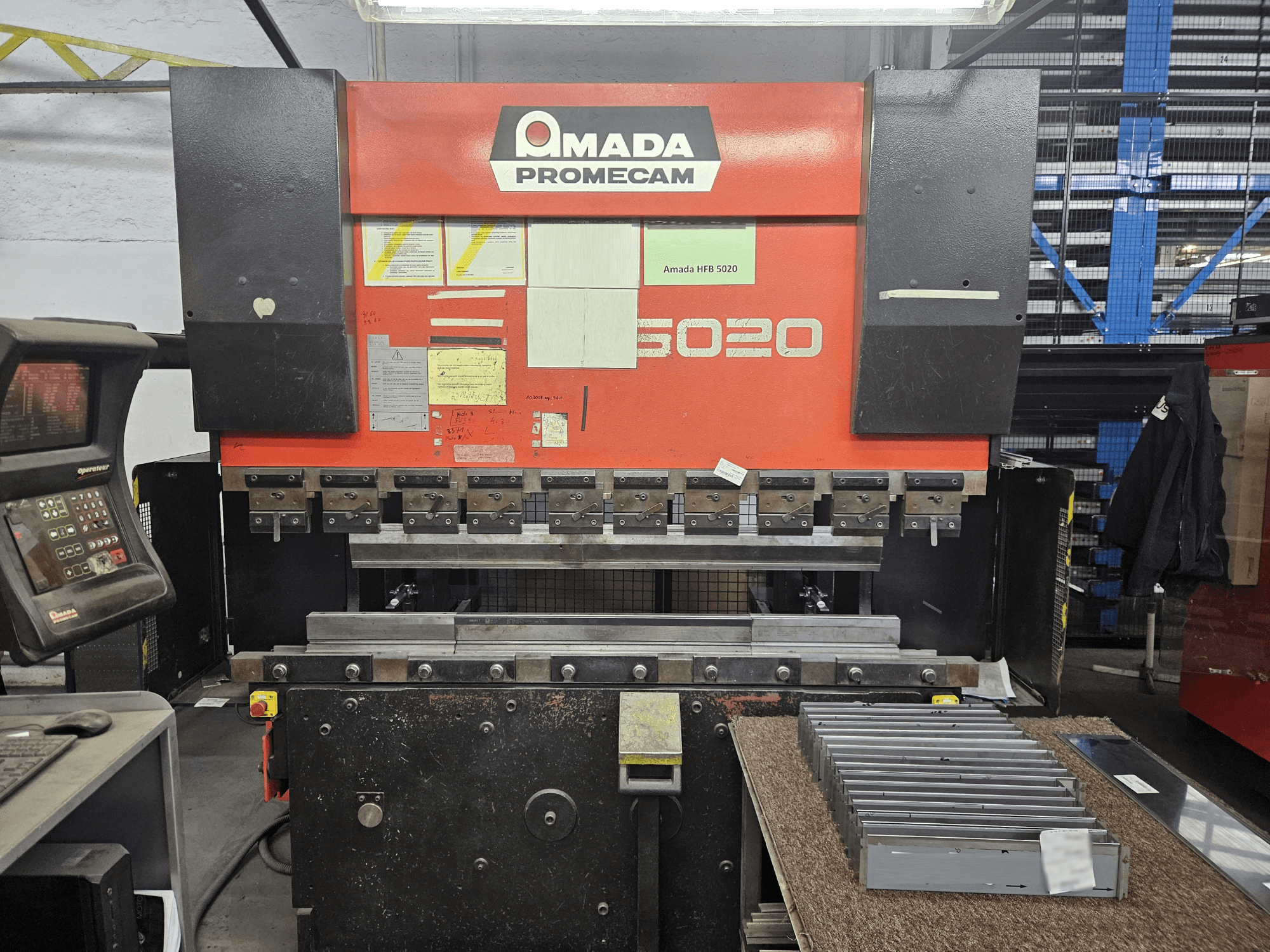 Pressa piegatrice Amada HFBO 5020 in vista frontale, con pannello di controllo e pezzi metallici impilati sulla superficie di lavoro.