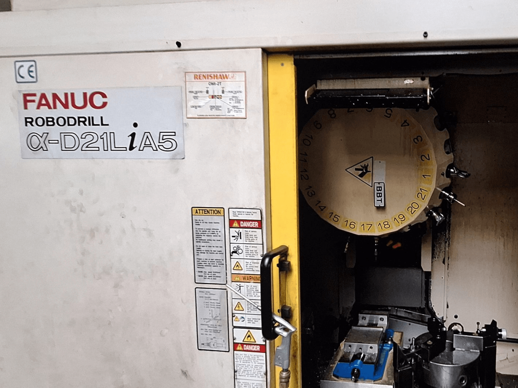 FANUC Robodrill α-D21LiA5, vista frontale. Mostra il carosello degli utensili e l'area di lavorazione con i morsetti e le avvertenze sullo sportello.