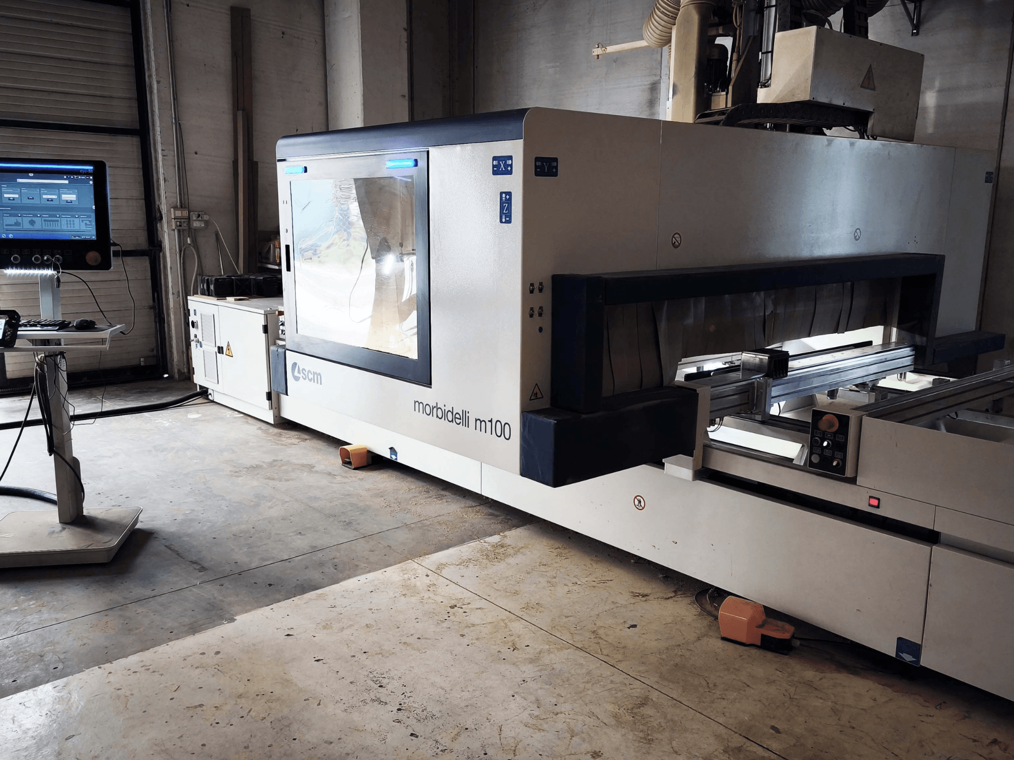 Centro di lavorazione del legno CNC SCM Morbidelli M 100 visto di lato, con in primo piano il pannello di controllo, l'area di lavoro e i dispositivi di sicurezza.
