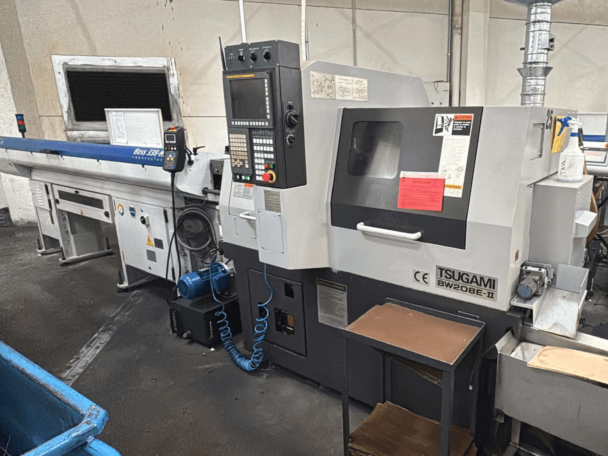 Macchina CNC Tsugami BW208E-II in un'officina, vista da un'angolazione frontale, con pannello di controllo ed etichette di sicurezza visibili.