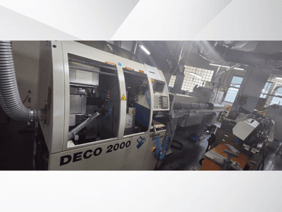 La macchina DECO 2000 CNC in officina, vista frontale, con i comandi e la configurazione di un pezzo lungo sulla destra.