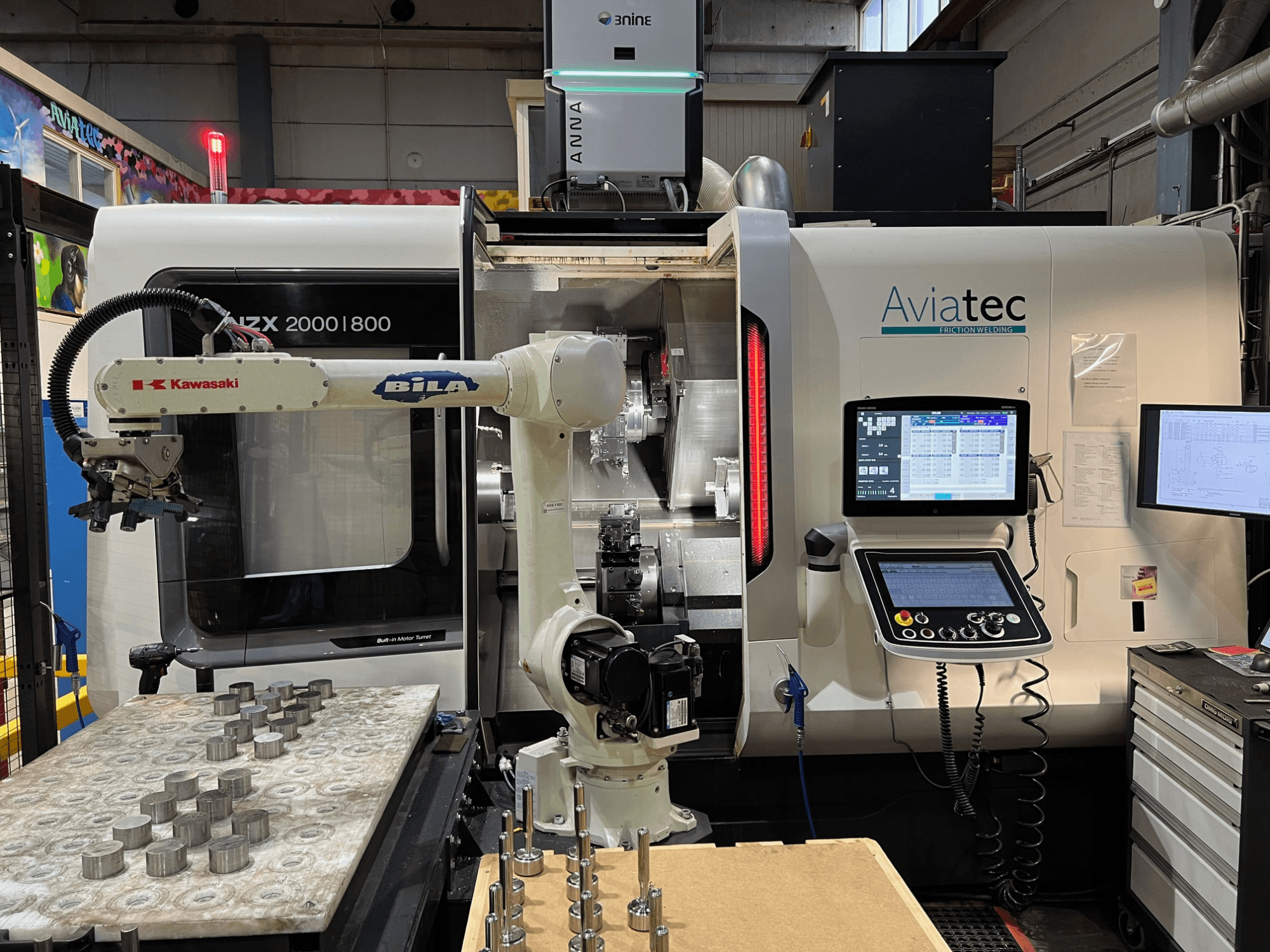 Tornio orizzontale DMG MORI NZX 2000/800SY con braccio robotico Kawasaki, pannello di controllo e componenti metallici sul piano di lavoro.