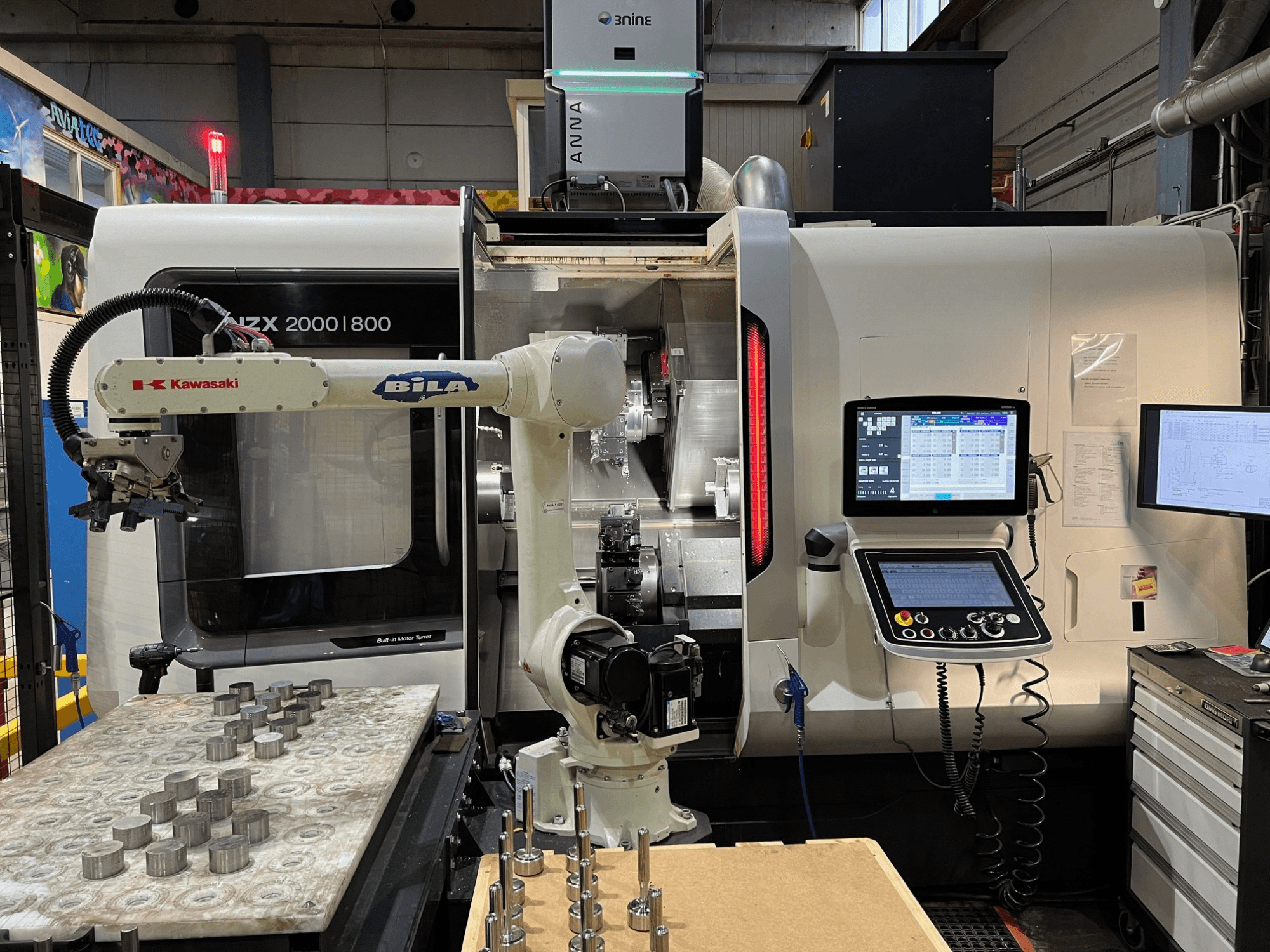 Tornio orizzontale DMG MORI NZX 2000/800SY in un'officina, con braccio robotico Kawasaki e comandi in vista.
