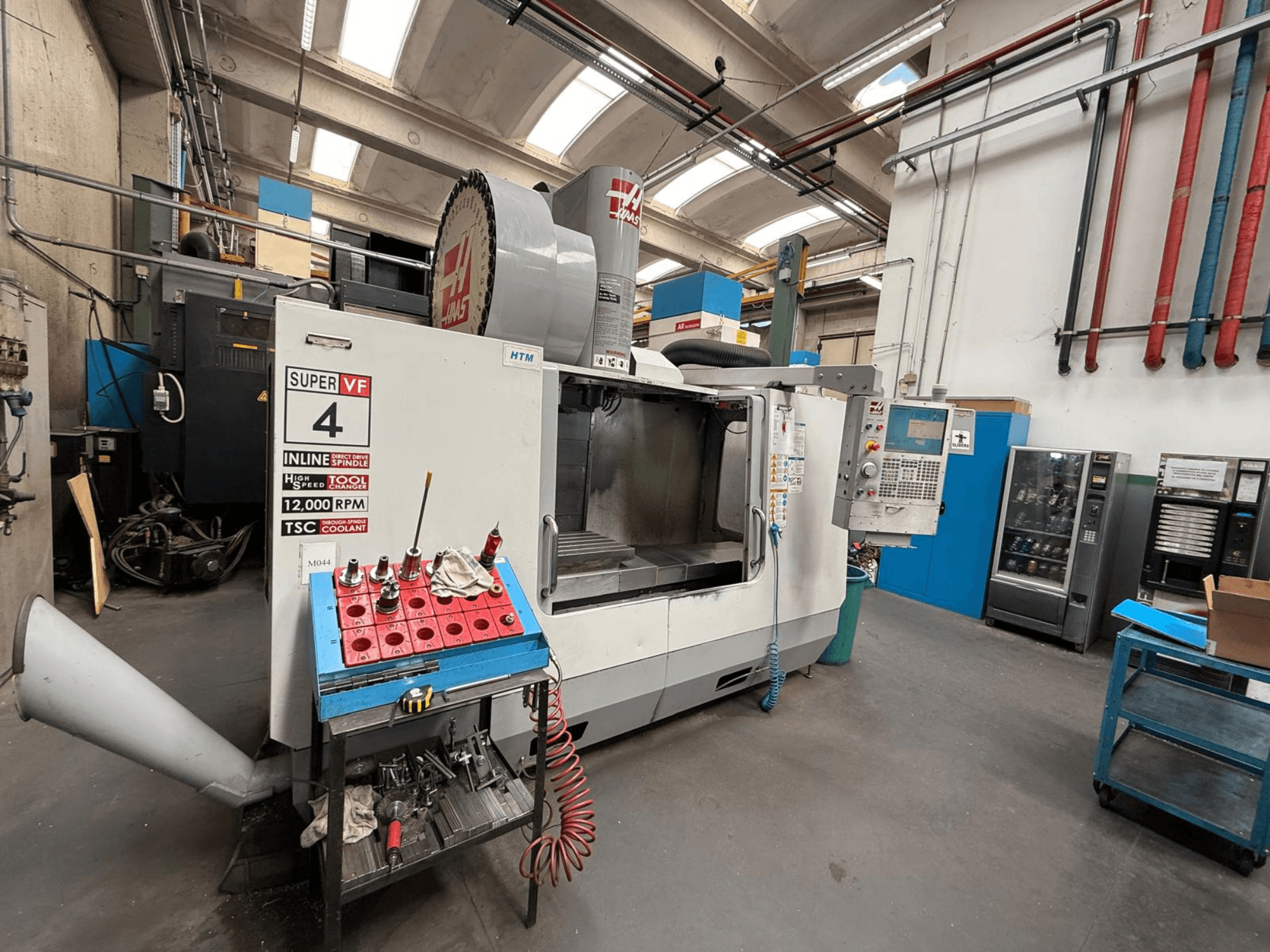 Fresatrice CNC HAAS Super VF 4 con pannello di controllo, mandrino da 12.000 giri/min e utensili su un rack di stoccaggio blu, vista laterale.