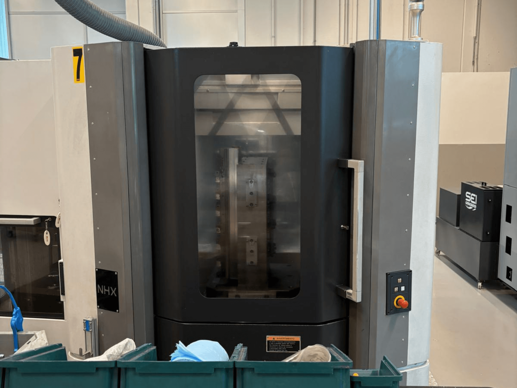 Macchina CNC NHX in vista frontale, con pannello di controllo e display chiaro; situata in un'officina con bidoni verdi nelle vicinanze.