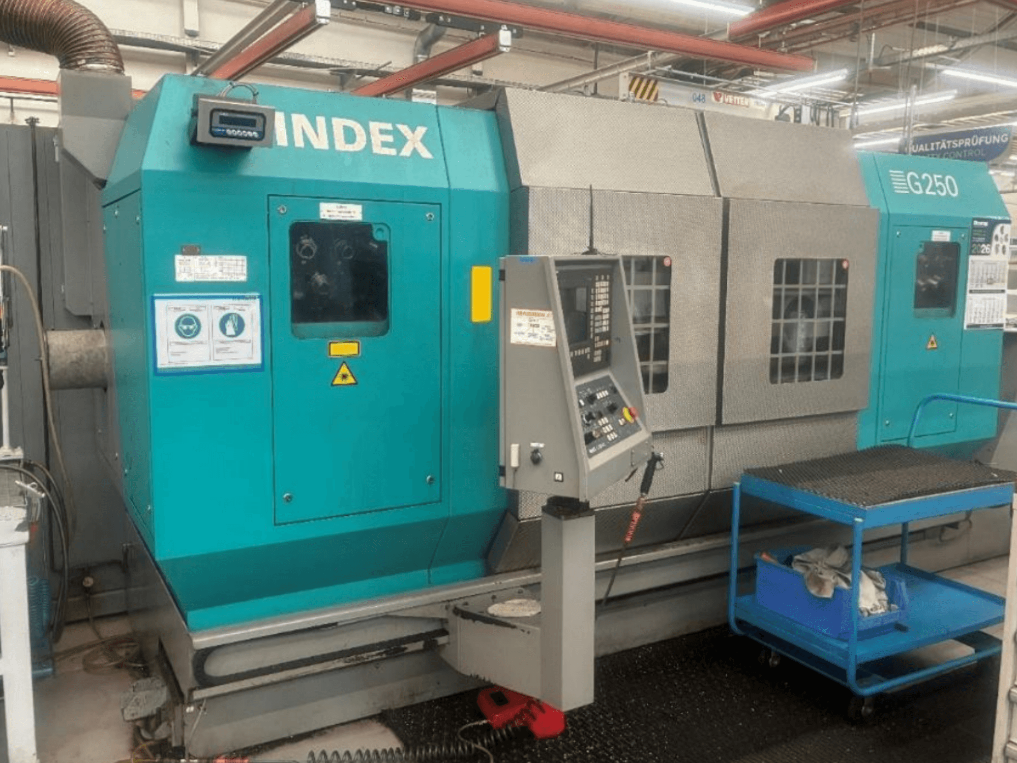 Tornio orizzontale INDEX G250, vista frontale che mostra il pannello di controllo e l'alloggiamento metallico, con etichette di sicurezza e un carrello blu.
