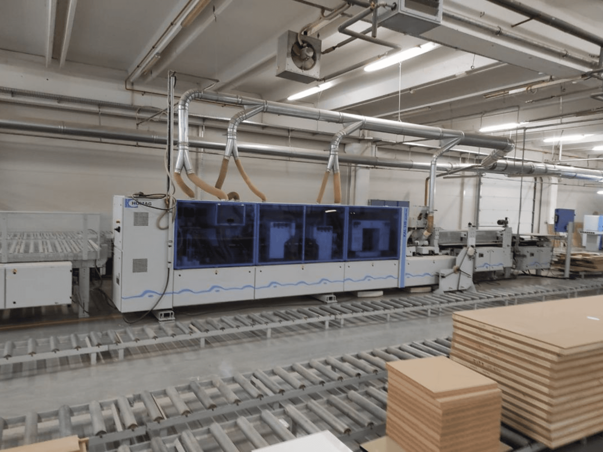 Macchina di produzione HOMAG in una fabbrica, vista frontale, con involucro blu e sistema di trasporto con fogli di compensato impilati.