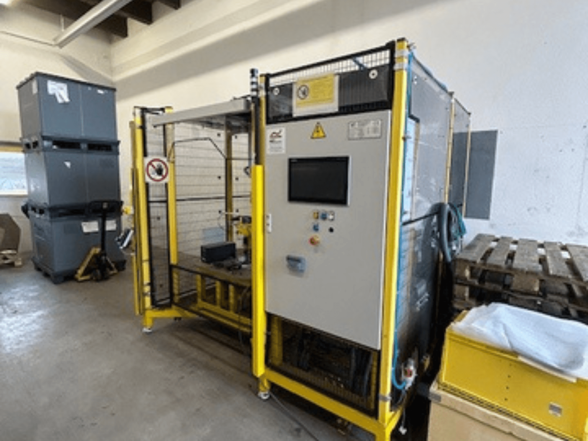 Macchina industriale con pannello di controllo e dispositivi di sicurezza, vista frontale; sullo sfondo telaio giallo, bidoni grigi e pallet.