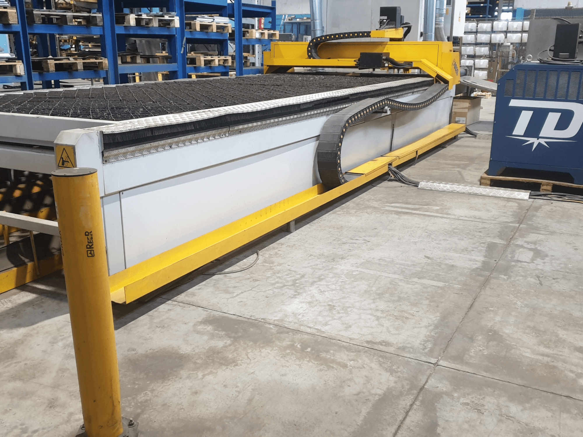 Macchina da taglio industriale con finiture gialle, vista frontale, con piano di lavoro e pannello di controllo testurizzati, collocata sul pavimento di una fabbrica.