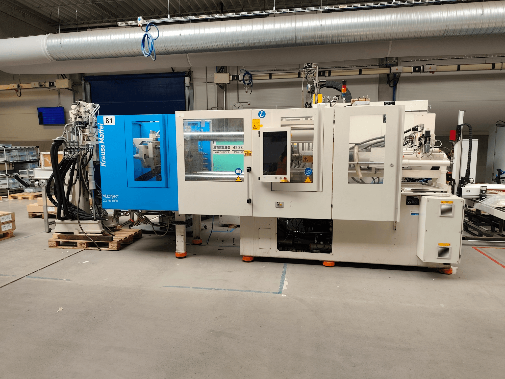 Macchina per stampaggio a iniezione idraulica Krauss Maffei 110-180-180 CX V, vista laterale, che mostra il corpo in blu e bianco, il pannello di controllo e i connettori.