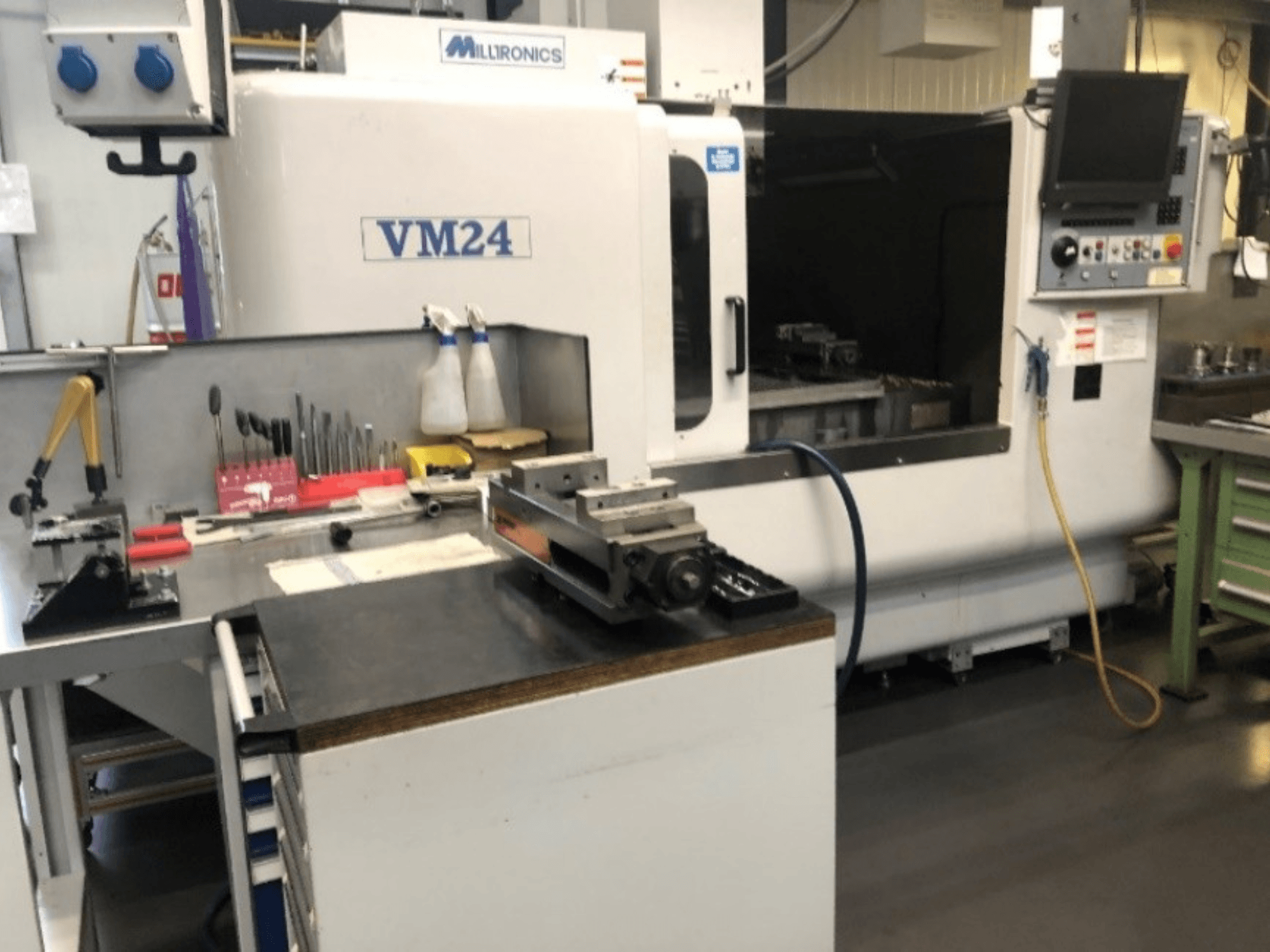 Area di lavoro con macchina CNC Milltronics VM24. Vista frontale con strumenti e attrezzature su un banco di lavoro.