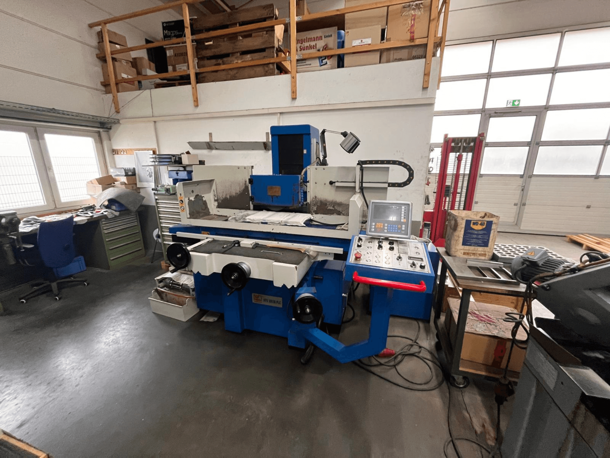 Smerigliatrice per superfici blu con pannello di controllo digitale, vista frontale, che mostra il disordine dell'area di lavoro e un armadietto degli attrezzi verde.