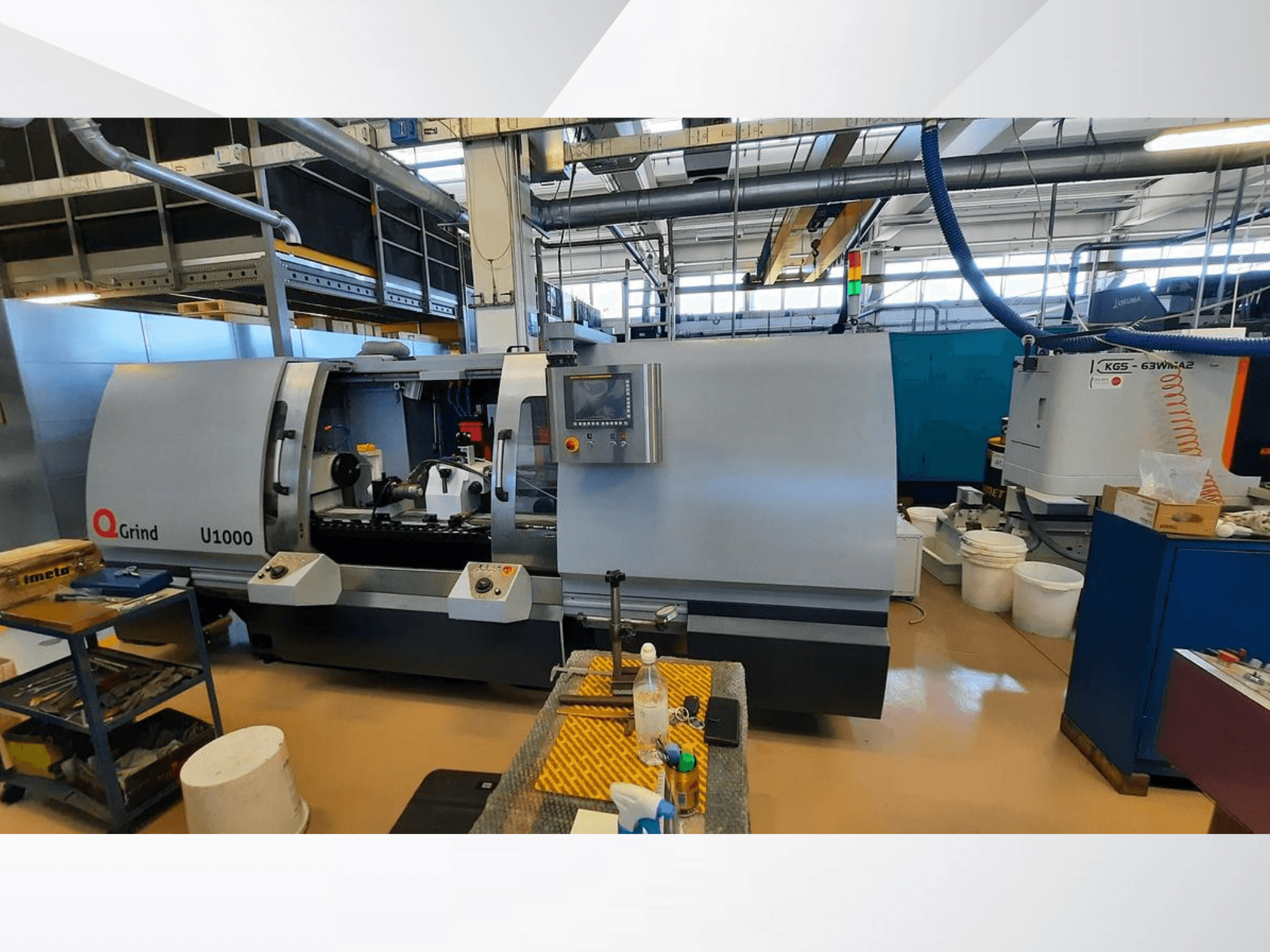 Macchina Q-Grind U1000 in un'officina, con vista frontale del pannello di controllo e dell'area di taglio aperta, circondata da attrezzature.