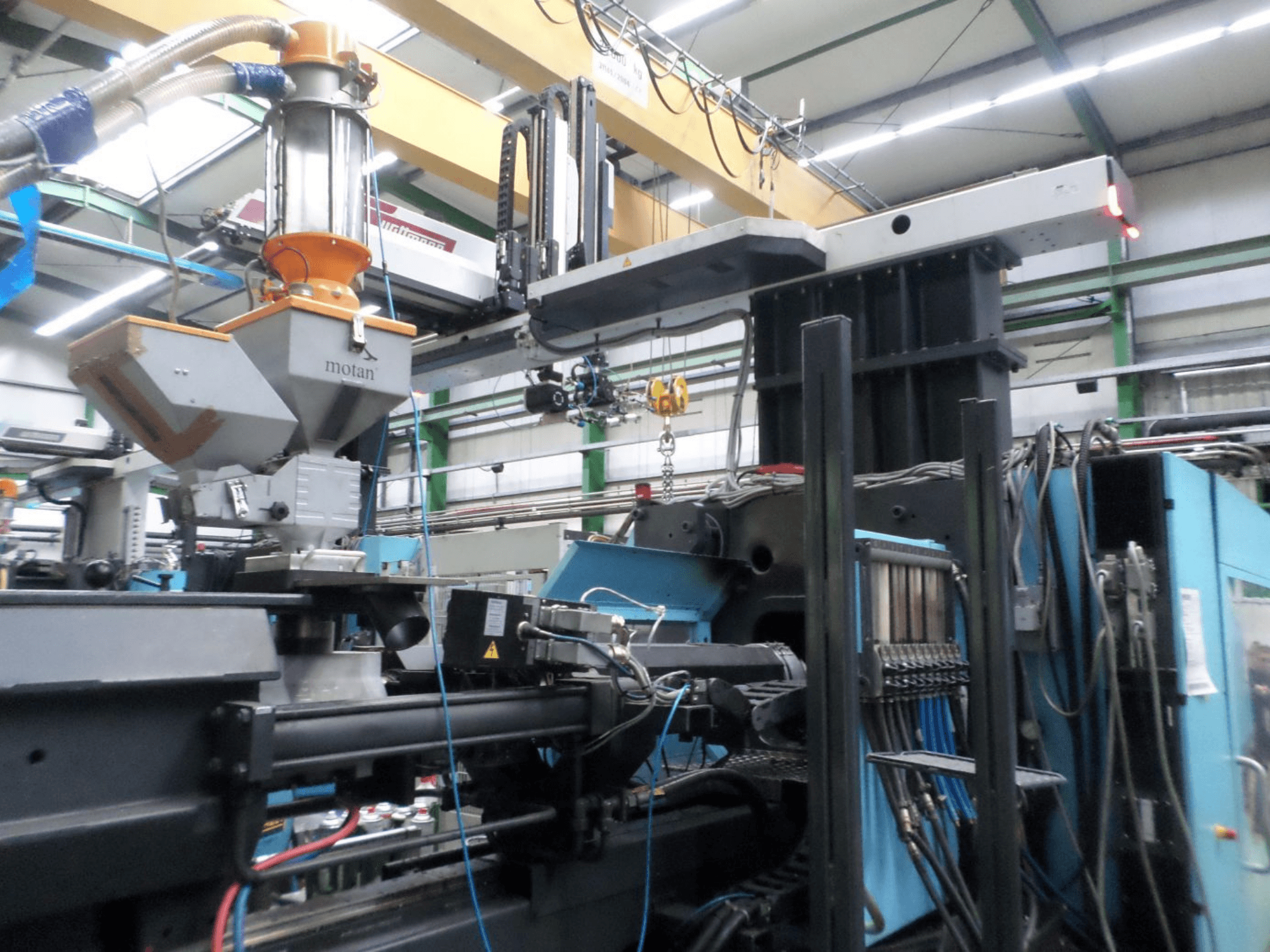 Macchina per lo stampaggio a iniezione idraulica DEMAG D 1300, vista laterale, che mostra la tramoggia, l’unità di controllo e i collegamenti in un ambiente di produzione.