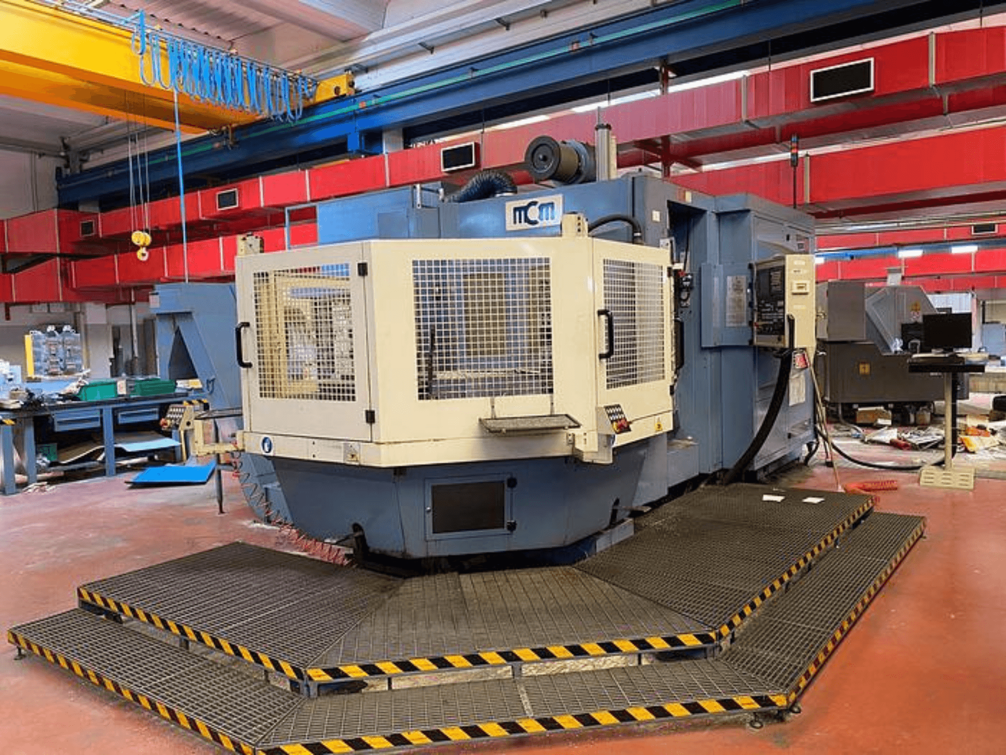 Macchina MCM CNC in una fabbrica, vista frontale con gabbia di protezione, bordi di sicurezza neri e gialli, circondata da attrezzature industriali.