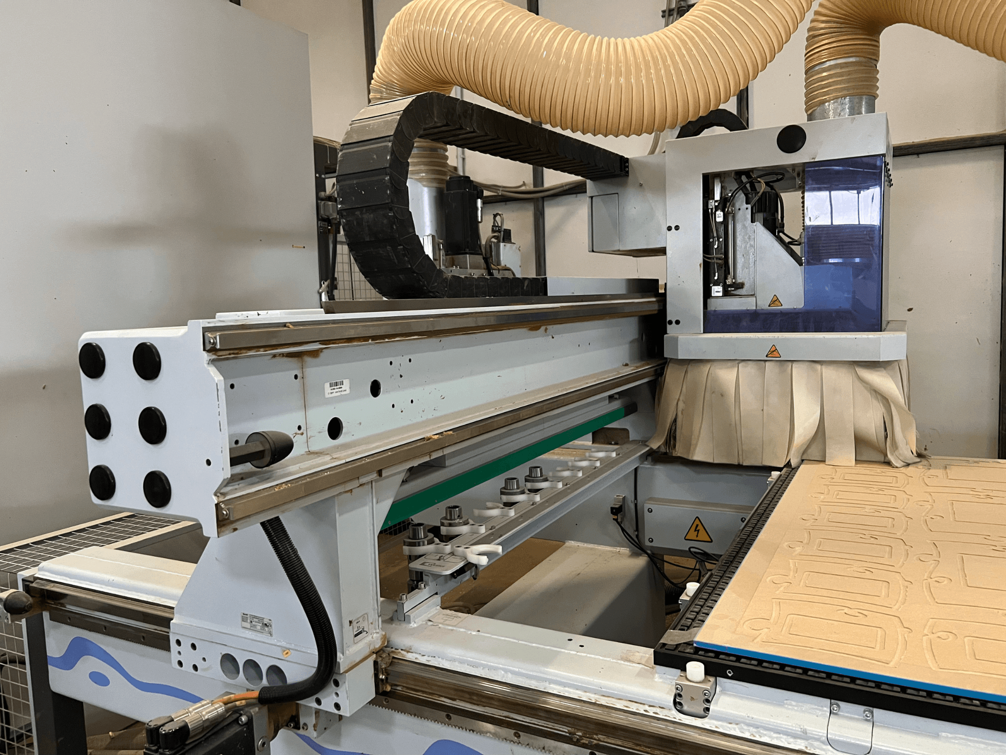 Vista laterale di un router CNC con pannello di controllo blu e argento, dotato di aspirazione della polvere e di un banco di taglio per il legno.