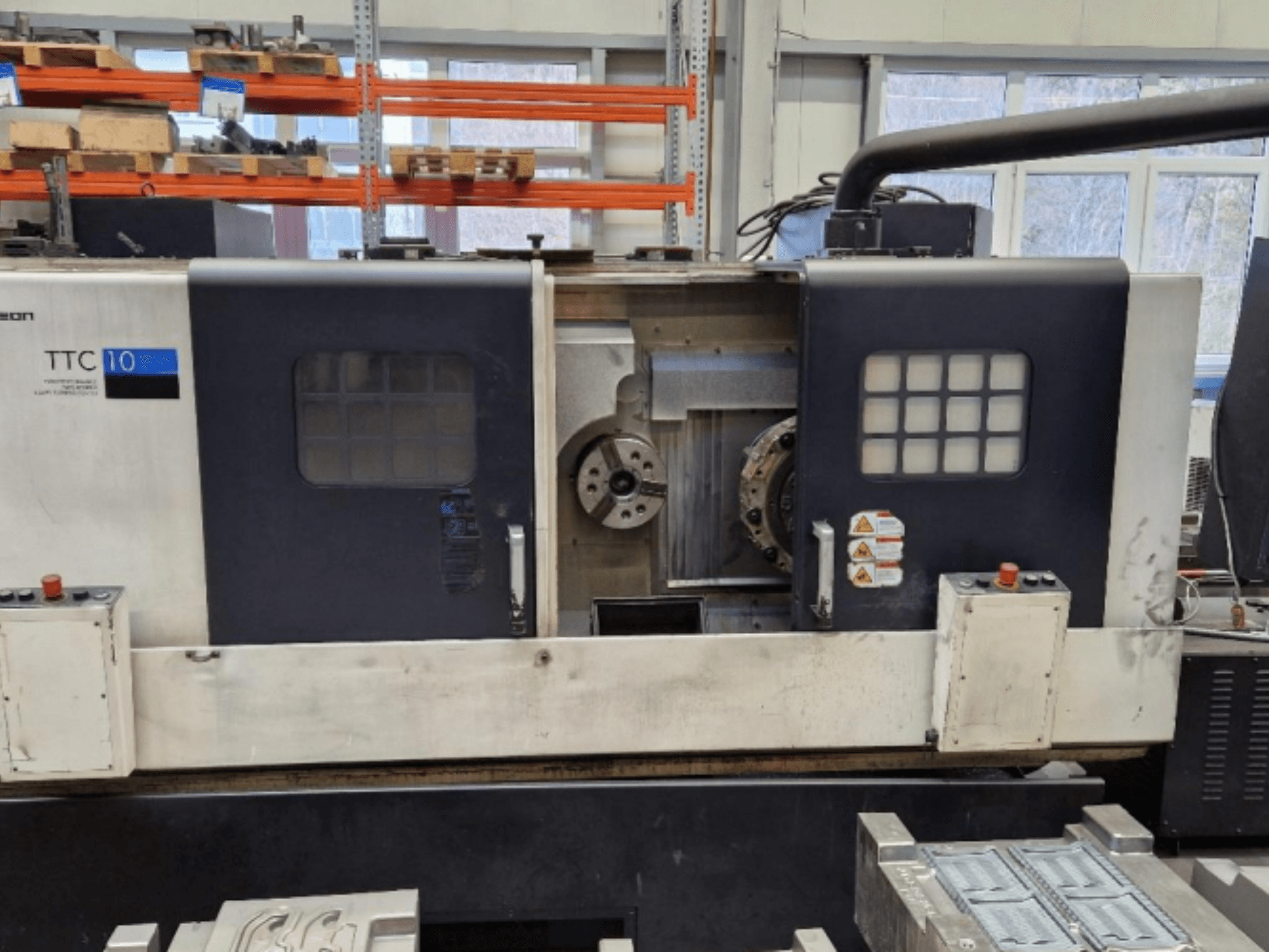 Vista frontale del tornio TTC 10, che mostra il pannello di controllo, i mandrini visibili e l'area di lavoro con i pezzi metallici.