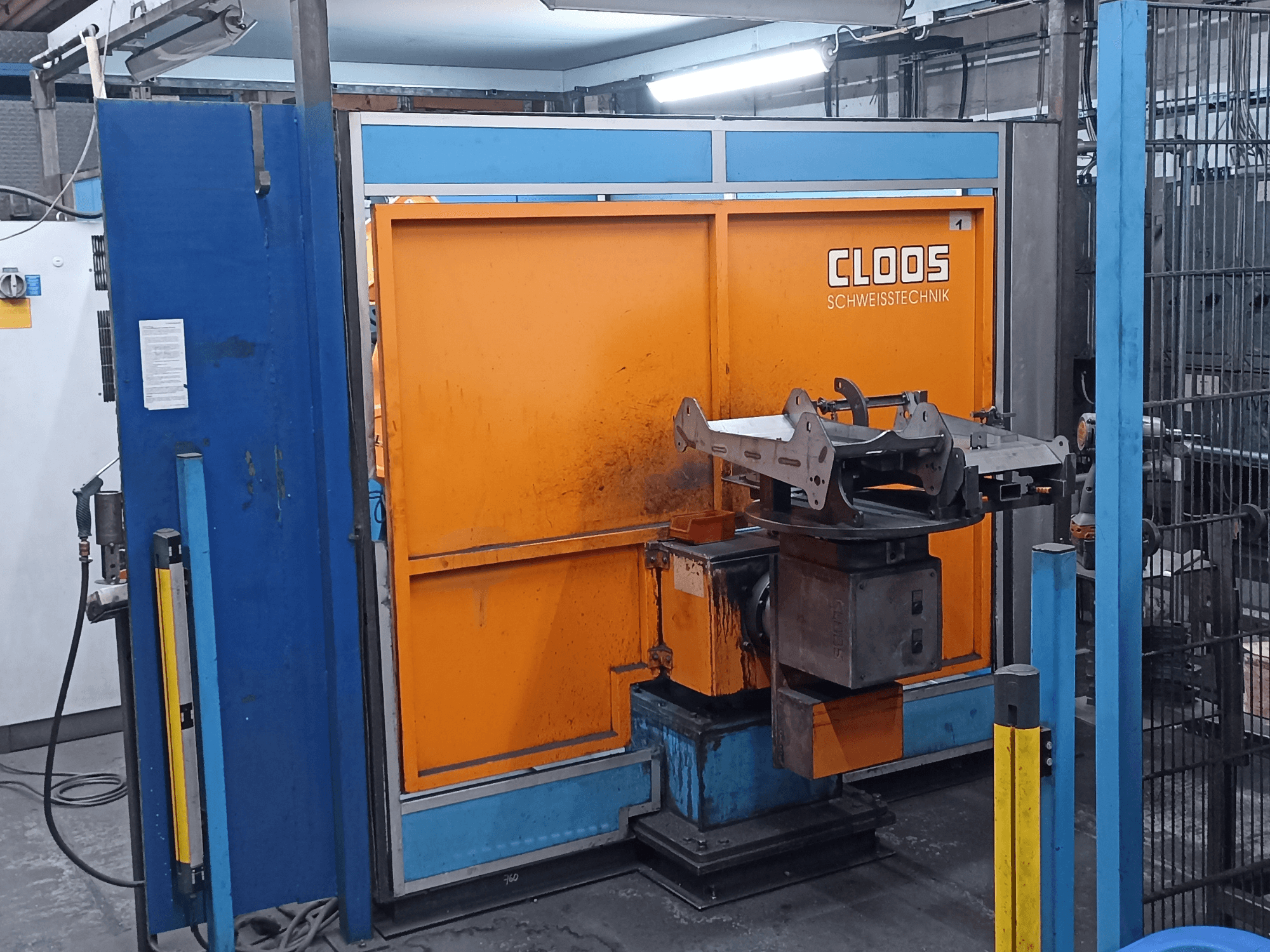 Robot di saldatura CLOOS Romat 320 in un'officina, vista frontale, con un pannello di controllo arancione e un pezzo da lavorare in metallo su una base girevole.