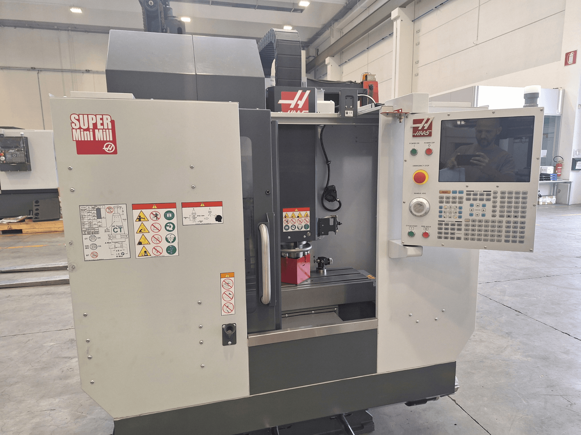 Vista frontale della fresatrice HAAS Super Mini Mill che mostra il pannello di controllo, le etichette di sicurezza e l'area di lavorazione con un pezzo in fase di posizionamento.