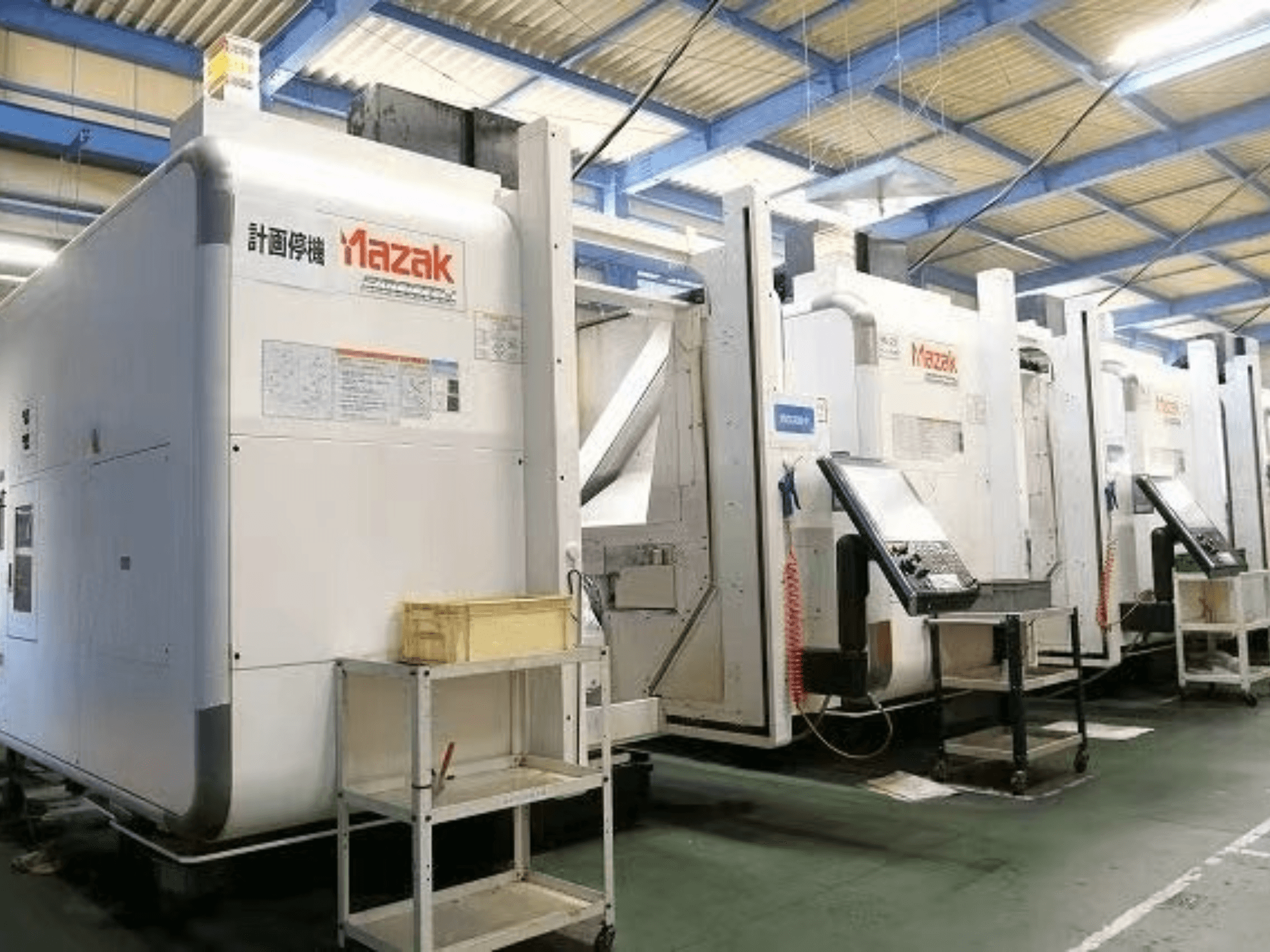 Macchine CNC Mazak in un ambiente di fabbrica, vista laterale che mostra pannelli di controllo ed etichette su corpi bianchi.