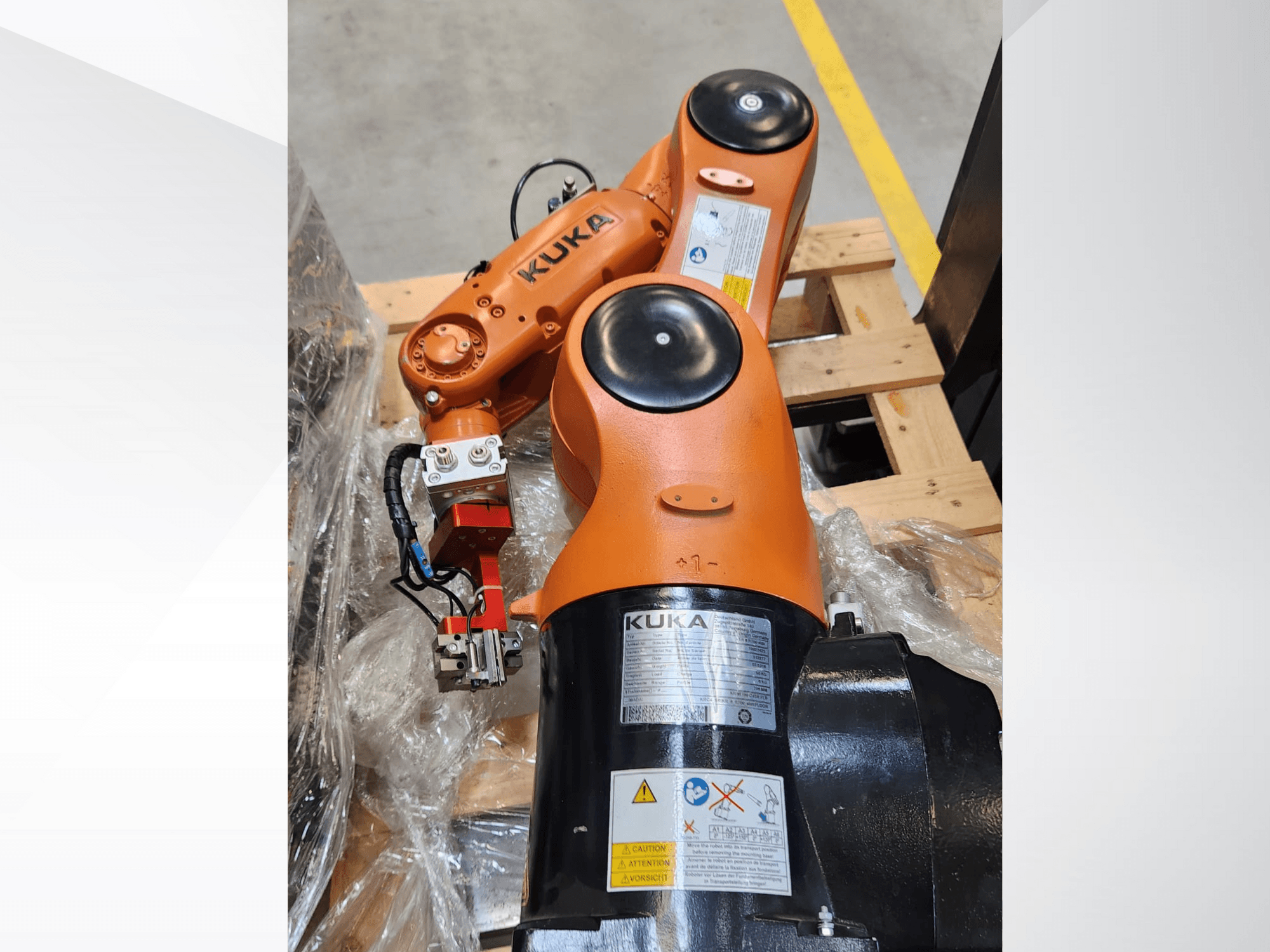 Braccio robotico KUKA con finitura arancione, vista laterale, con pinza e cablaggio di controllo su un pallet di legno.