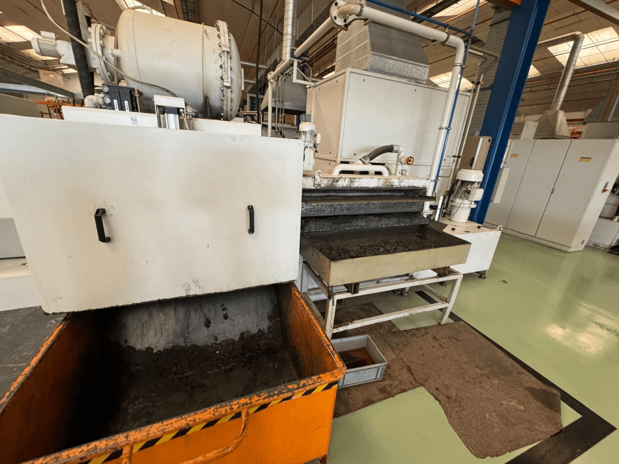 Apparecchiatura di lavorazione industriale con un contenitore di rifiuti arancione, vista frontale; contiene un motore, nastri trasportatori e un'area di manutenzione.